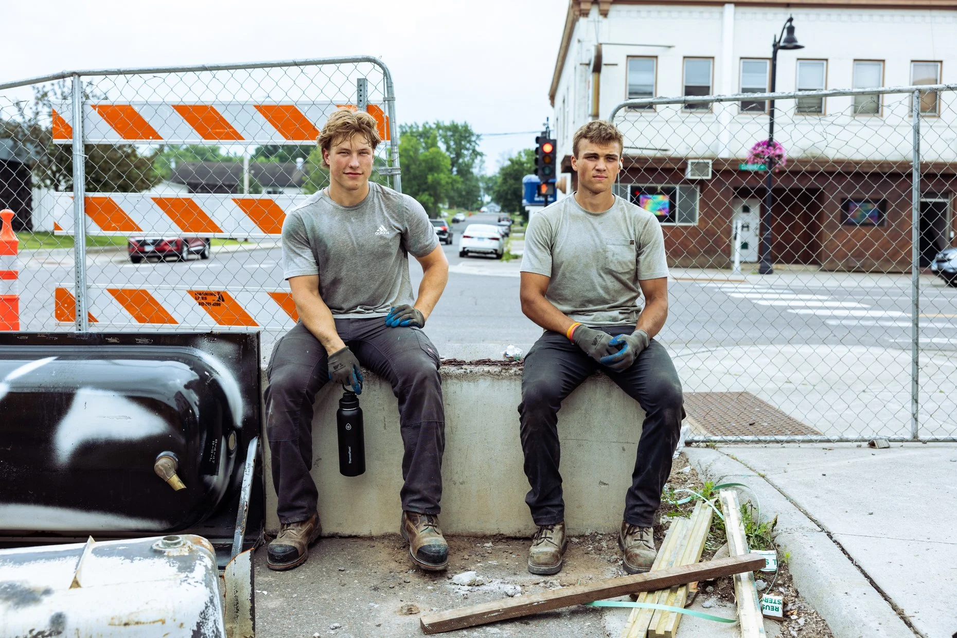 Two young men sit on a concrete barrier in front of a chain-link fence with construction barricades behind them. Both are wearing work gloves and casual work clothes, with a water bottle and tools nearby. An old vehicle part is in the foreground.