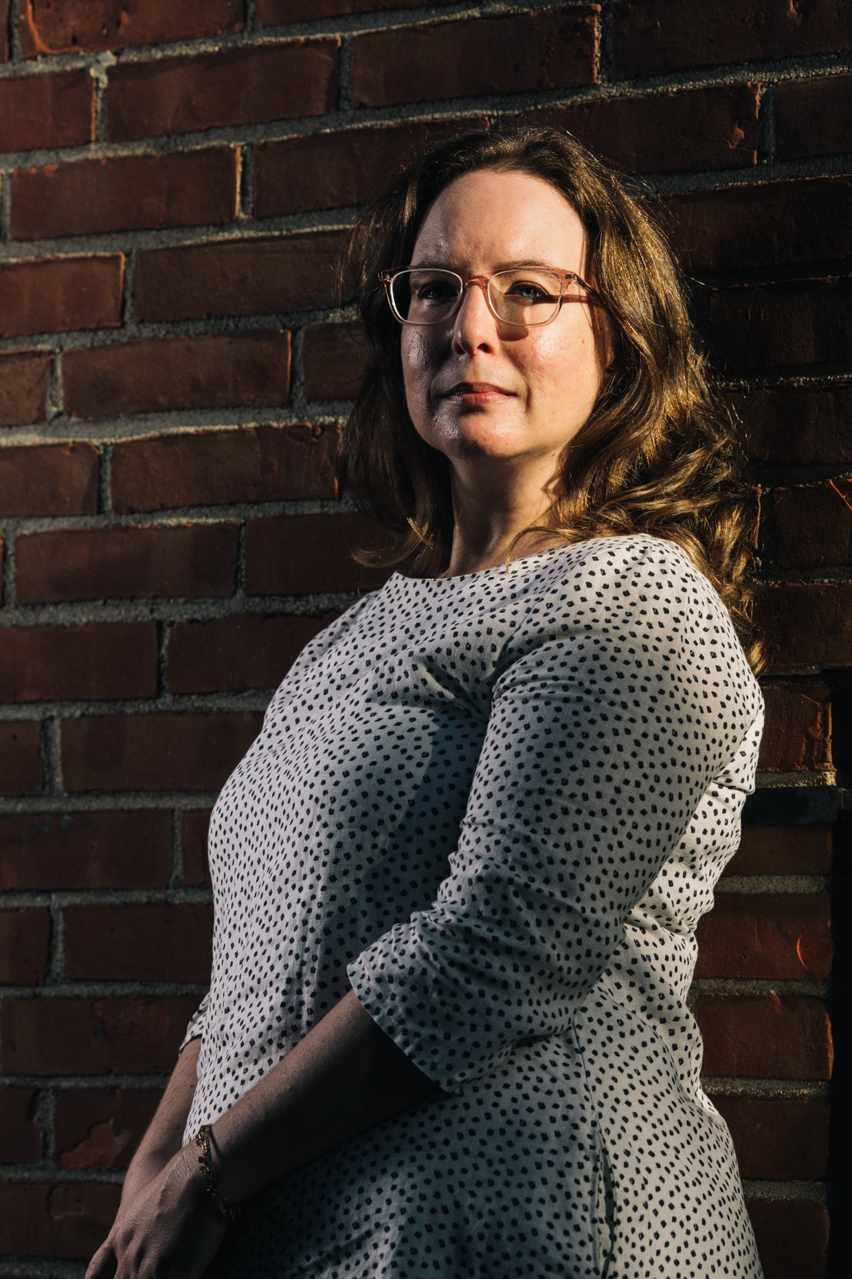 A woman with glasses and wavy hair standing against a brick wall, wearing a white polka dot dress.