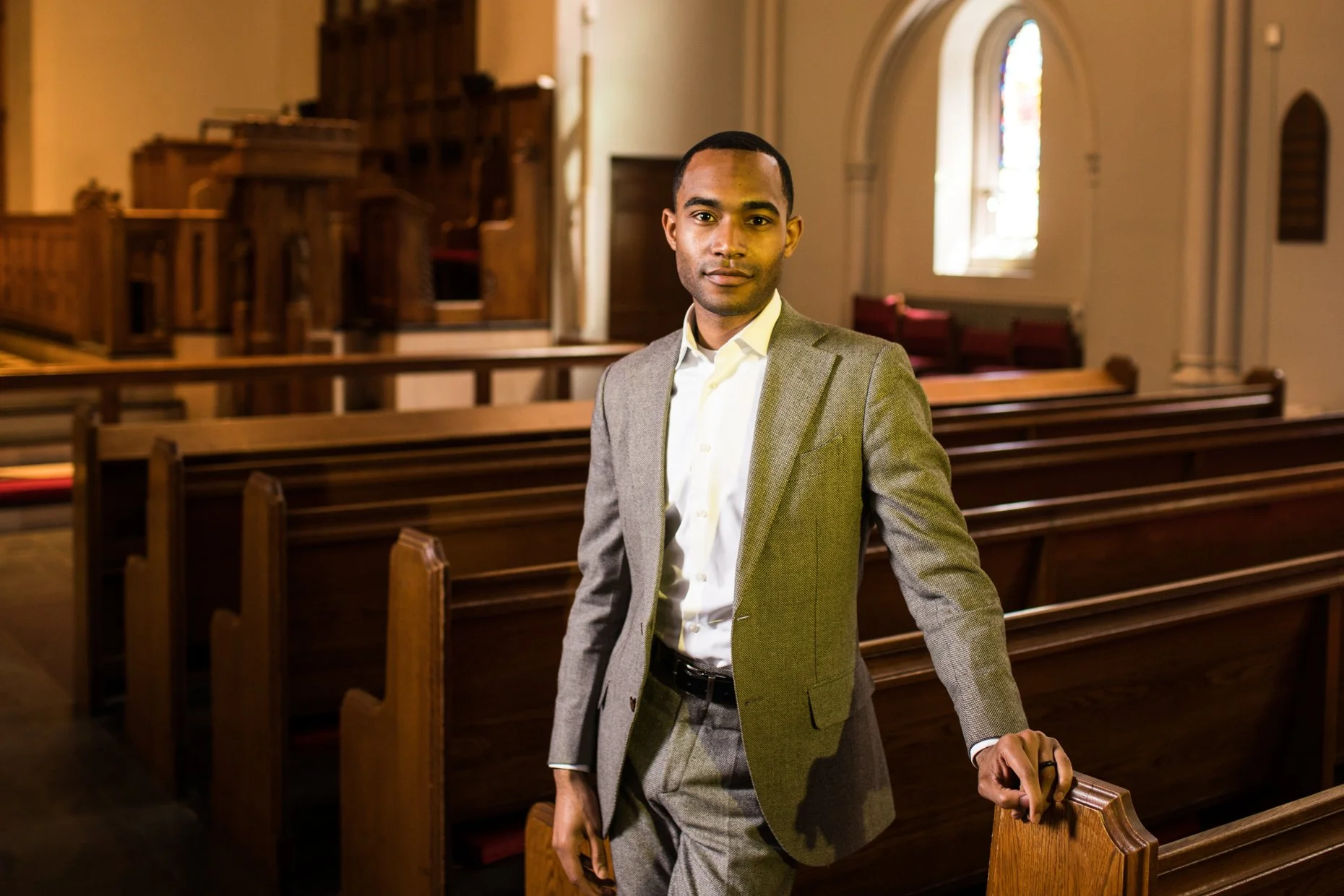 A man in a gray suit and white shirt standing in a church with wooden pews and stained glass windows.