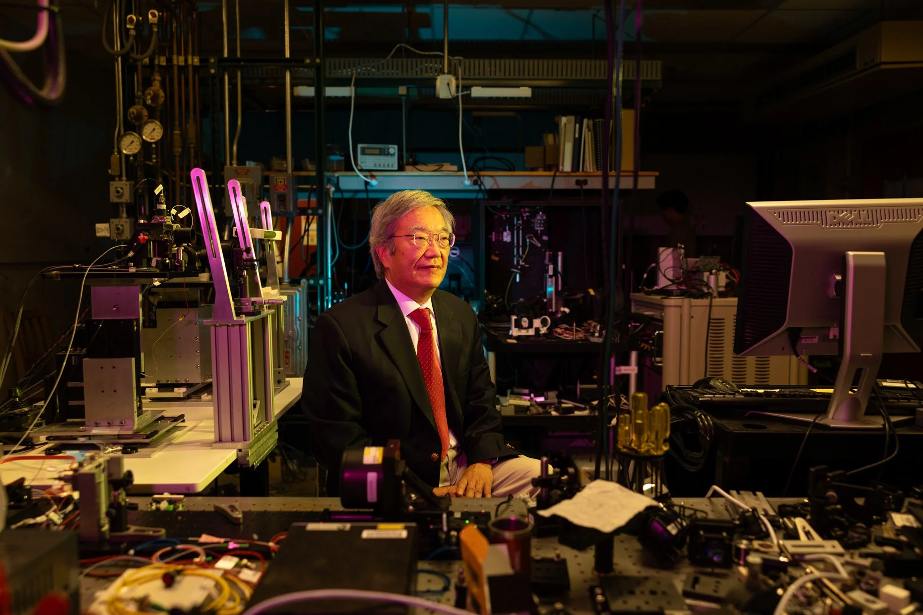 A man in a suit and glasses sitting in a laboratory filled with scientific equipment, computers, and various technical instruments.