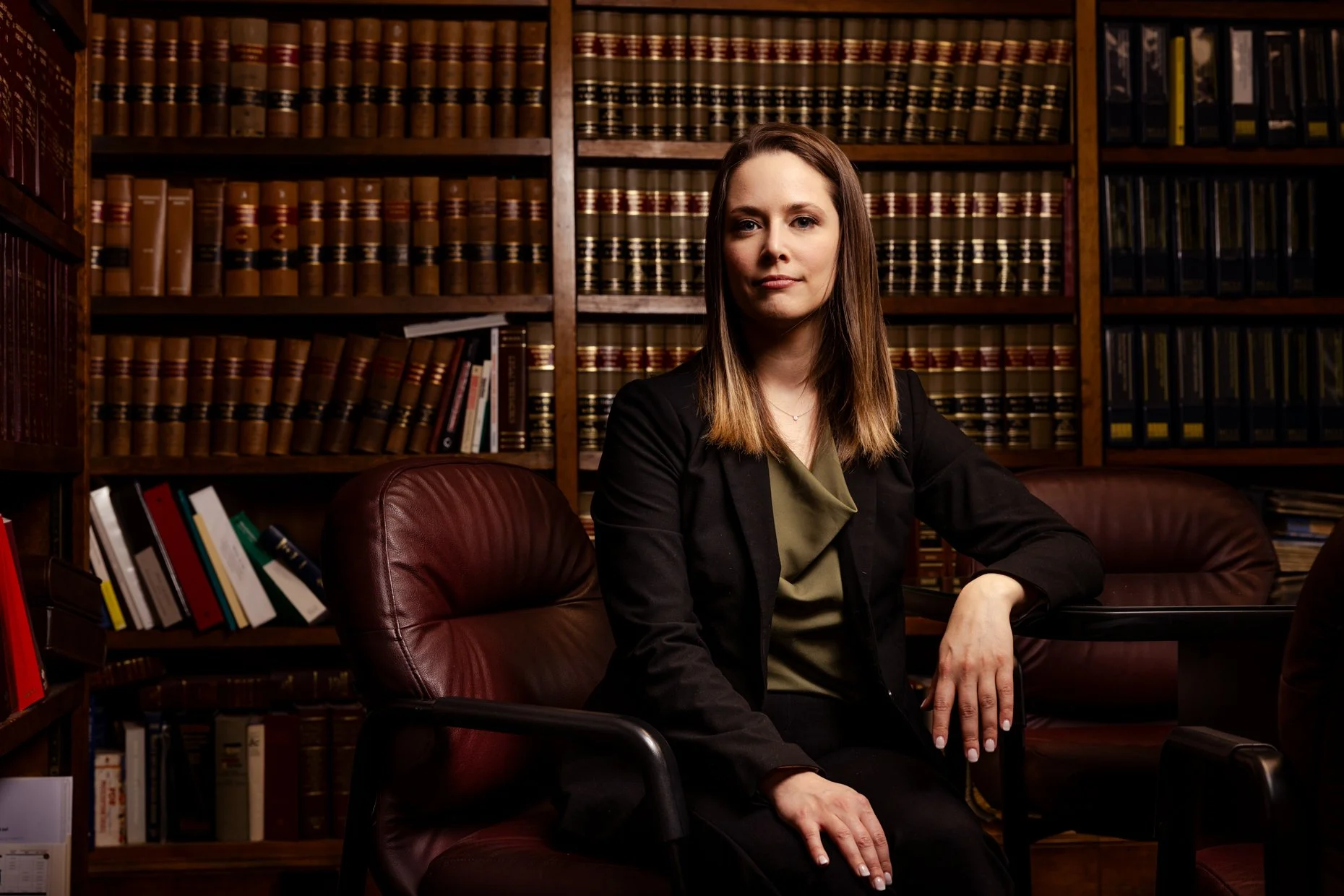 A woman with long brown hair dressed in a black blazer and green blouse seated in a leather chair at a wooden table in a law library, with bookshelves filled with legal books in the background.