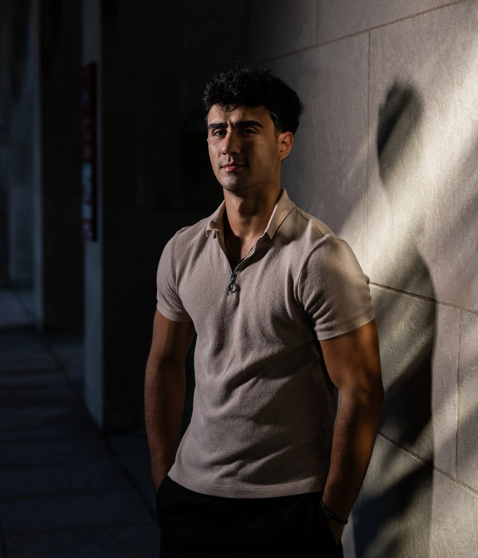 A young man with dark, curly hair standing against a textured wall. He is wearing a beige zip-up shirt and a bracelet on his left wrist. The lighting creates a shadow of his profile on the wall.