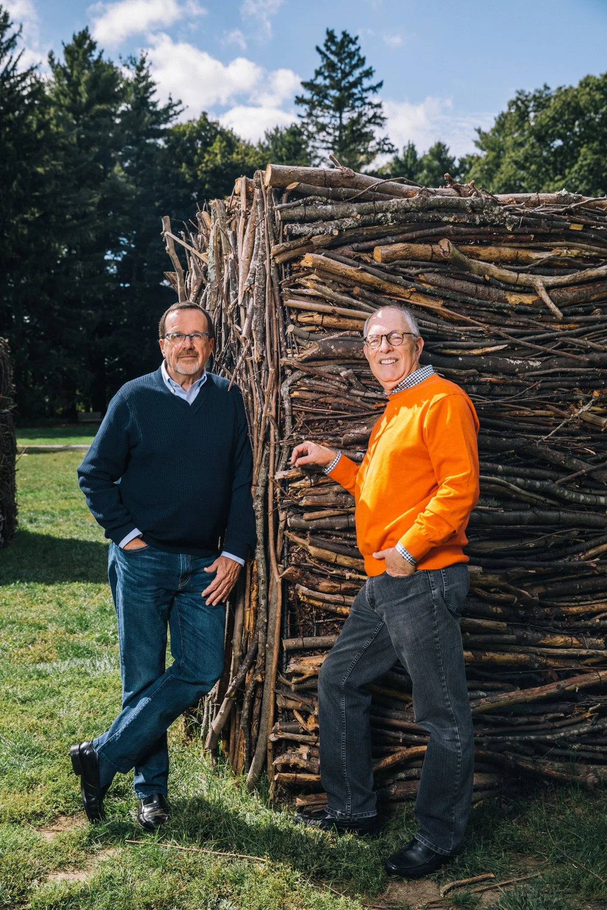 Two middle-aged men standing outdoors next to a large structure made of stacked wooden sticks or logs, with a background of trees and a partly cloudy sky.