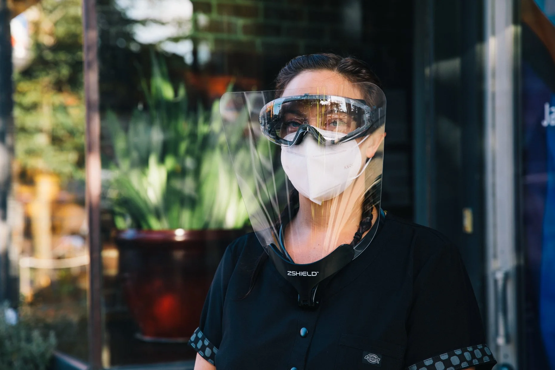 A woman wearing a white KN95 face mask, protective goggles, and a transparent face shield. She has dark hair in braids and is wearing a black shirt with patterned sleeves. She appears to be standing outdoors with greenery and a building in the backgr