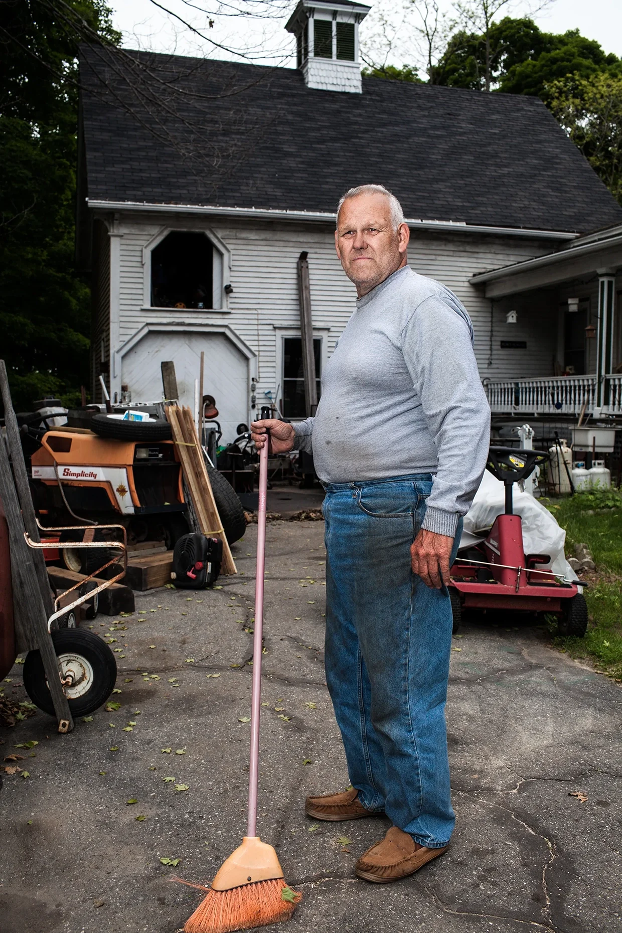 An elderly man standing outdoors in front of a house, holding a pink broom, wearing a gray sweatshirt, blue jeans, and brown shoes, with various tools and equipment around him.