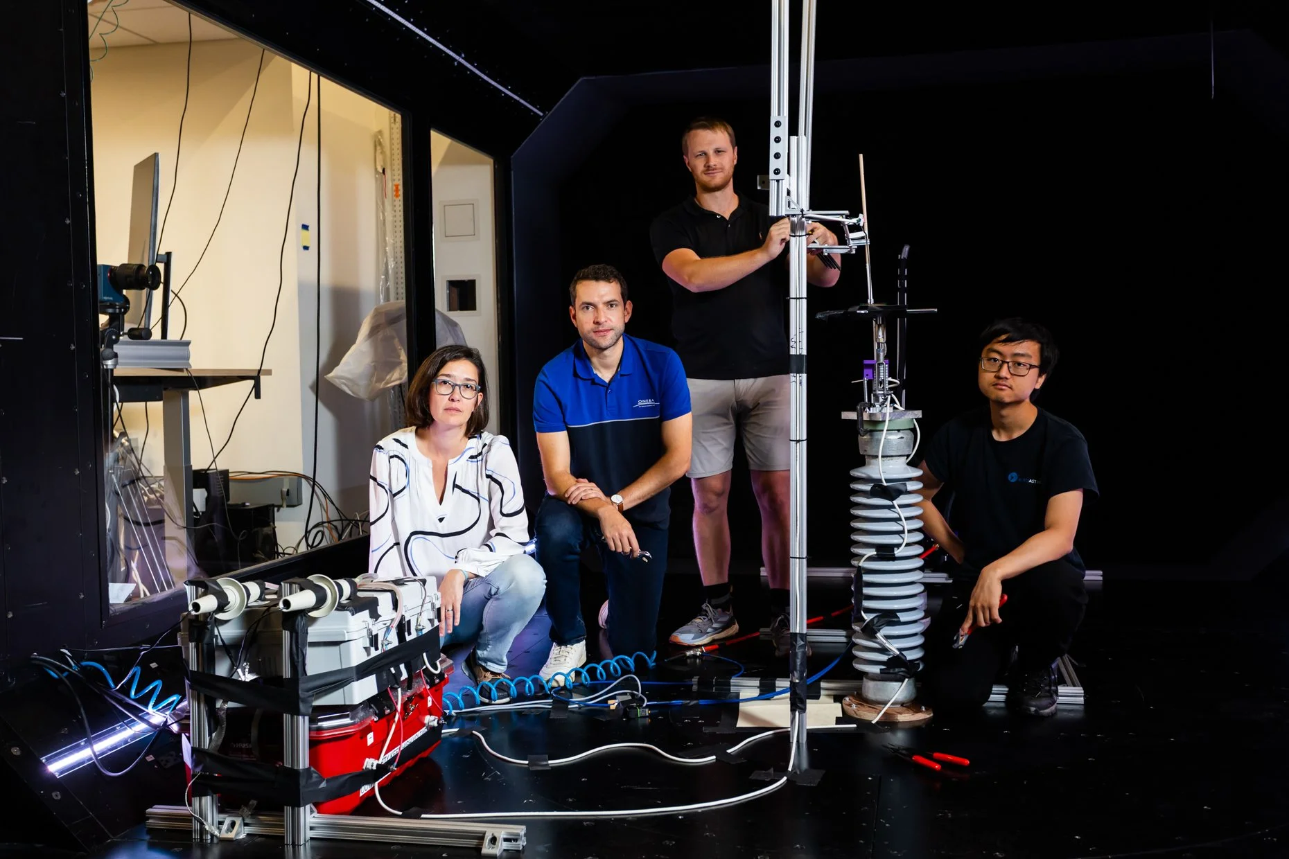 Group of four scientists working in a lab with scientific equipment, including a vertical metal structure, wires, and computer devices, in a darkened room.