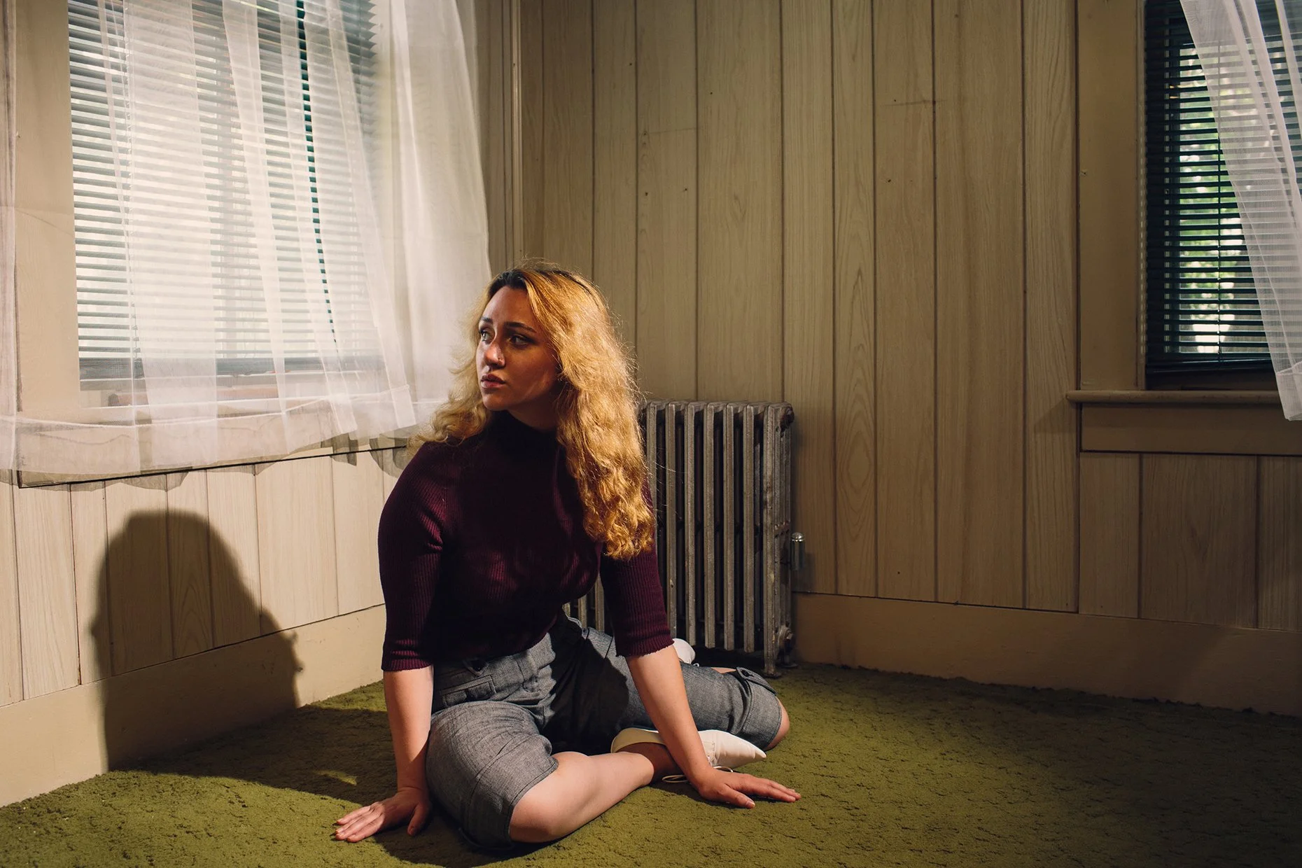 A woman with curly blonde hair, wearing a black top and gray pants, sits on a green carpeted floor in a room with wood-paneled walls. She is looking to her left with a contemplative expression. The room has a window with semi-sheer curtains, a radiat