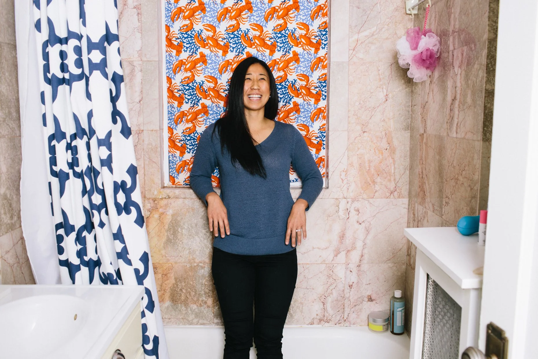 Woman smiling in a bathroom with pink marble walls, a shower curtain with a blue and white pattern, a backsplash with orange and blue coral and lobster design, and various toiletries on a small white shelf.