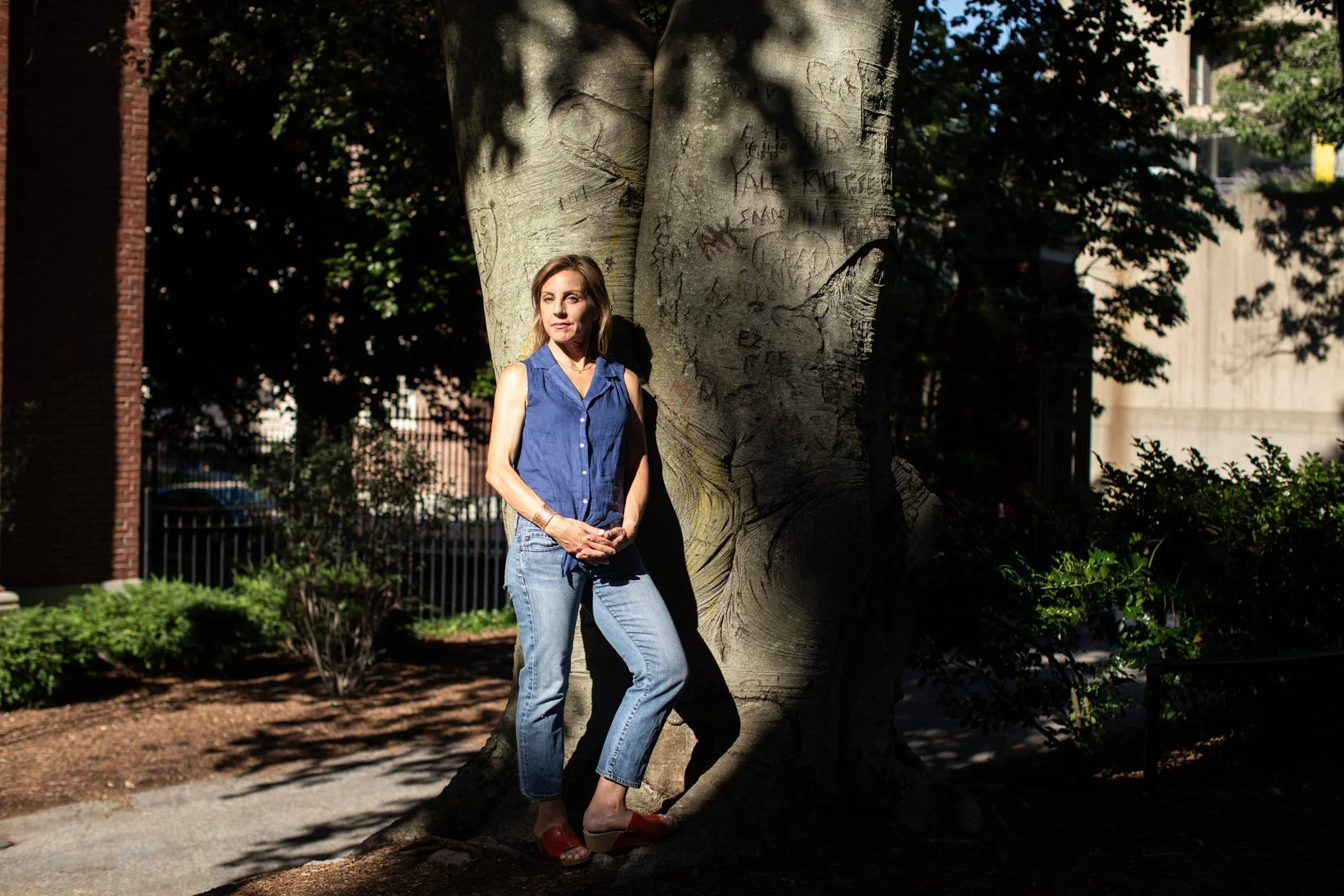 A woman in a blue sleeveless shirt and light blue jeans stands against a large tree in a park during daytime. She is standing with her arms loosely clasped in front of her, looking slightly to the side. Shadows of leaves are cast on her face and the 