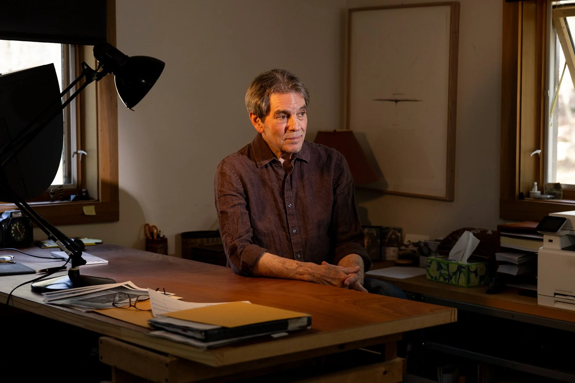An older man with gray hair sitting at a large wooden desk in a home office, with windows behind him, surrounded by papers, a laptop, and office supplies.