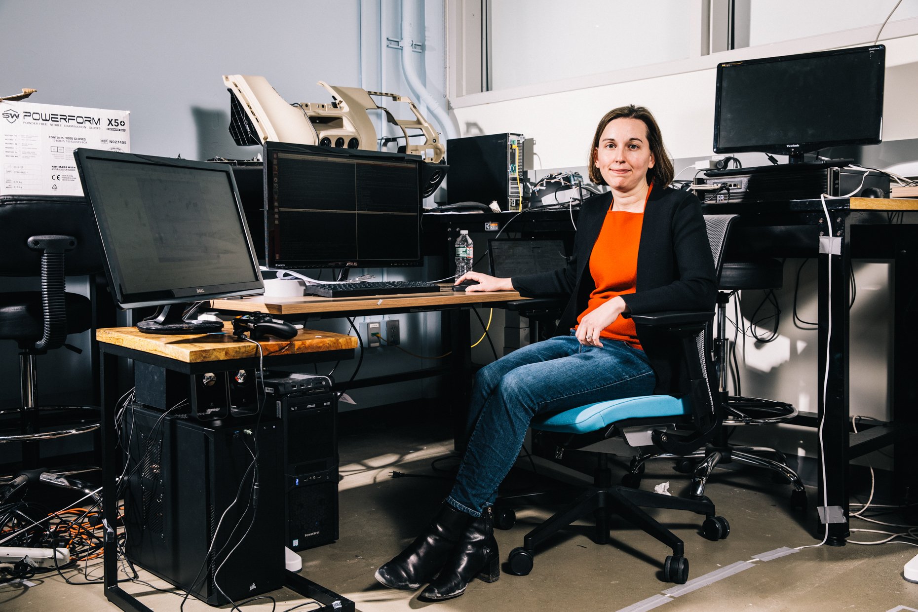 A woman sitting at a computer workstation with multiple monitors and electronic equipment in a laboratory or tech workspace.