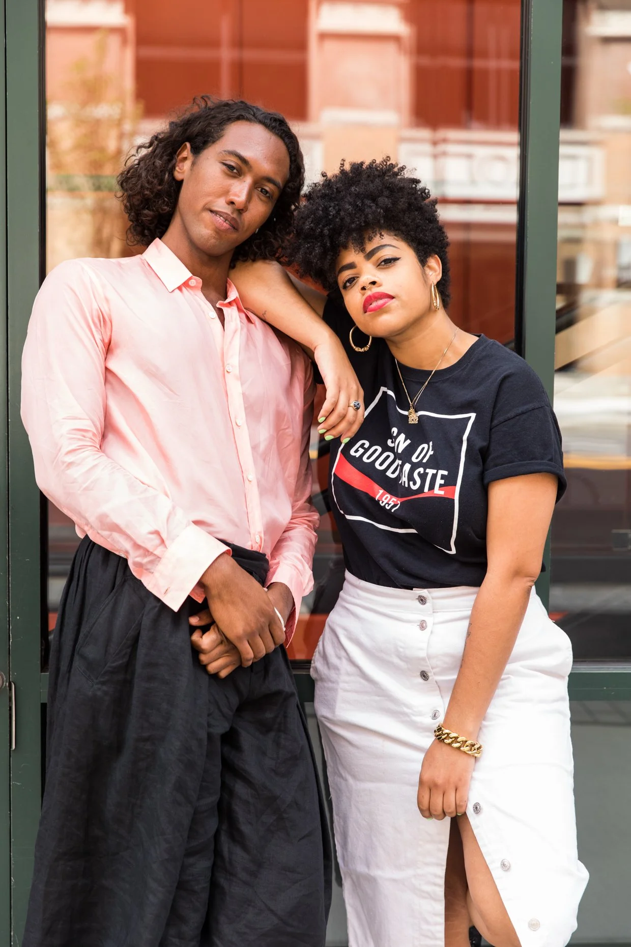 Two people standing in front of a window with a city street background. The person on the left has long curly hair, wearing a light pink shirt and dark pants. The person on the right has short curly hair, wearing a black T-shirt with white and red de