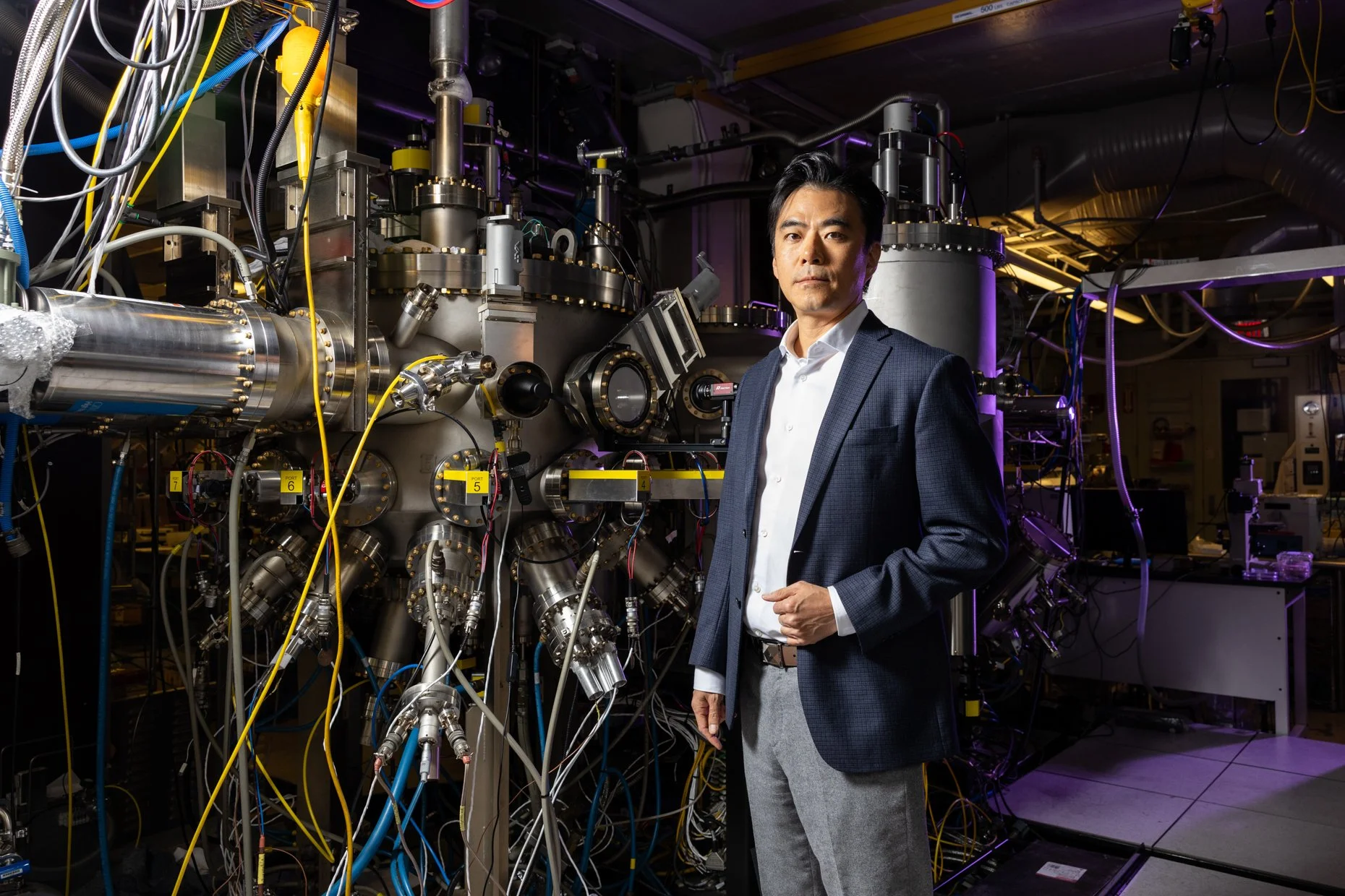 A man in a suit stands in front of complex scientific equipment with numerous pipes, wires, and metallic components in a laboratory setting.