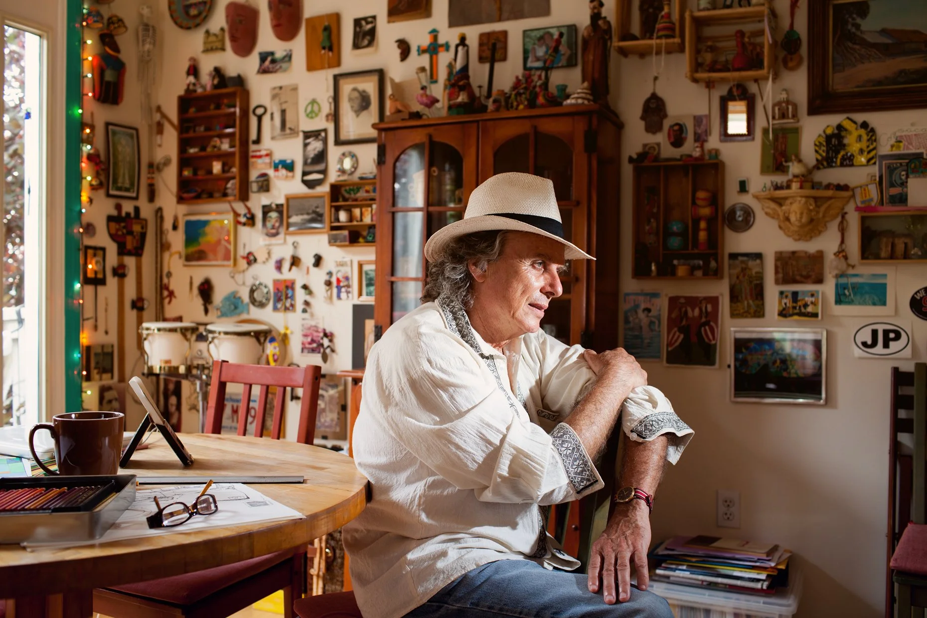 An older man wearing a light-colored shirt and a white hat with a black band sits at a wooden table, resting his arms on the table in a room decorated with numerous colorful artwork, masks, and trinkets on the walls. A window with string lights is vi