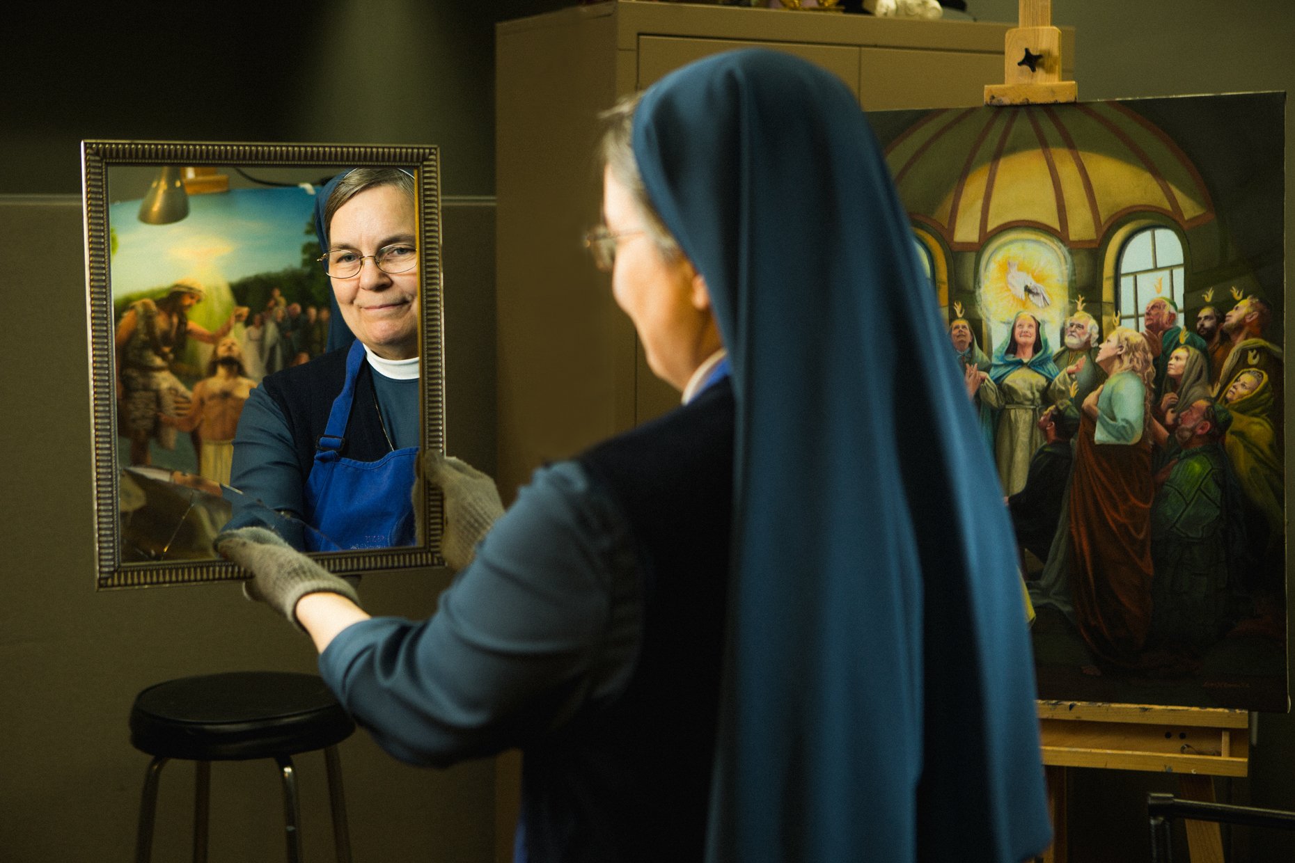 A woman dressed as a nun is painting a portrait of a person in a mirror. Behind her, there is a colorful painting of a religious scene.