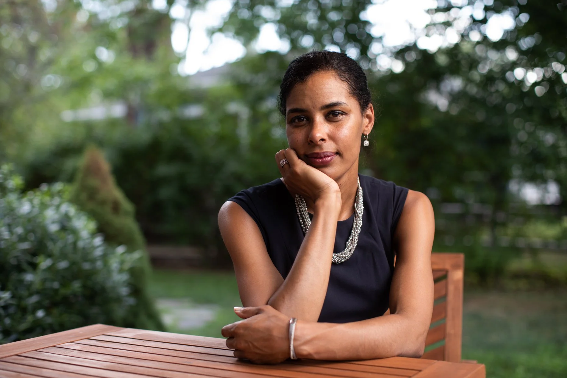 A woman sitting outdoors at a wooden table, resting her chin on her hand, with a thoughtful expression. She is wearing a black sleeveless top, pearl jewelry, and earrings, with blurred green trees and bushes in the background.