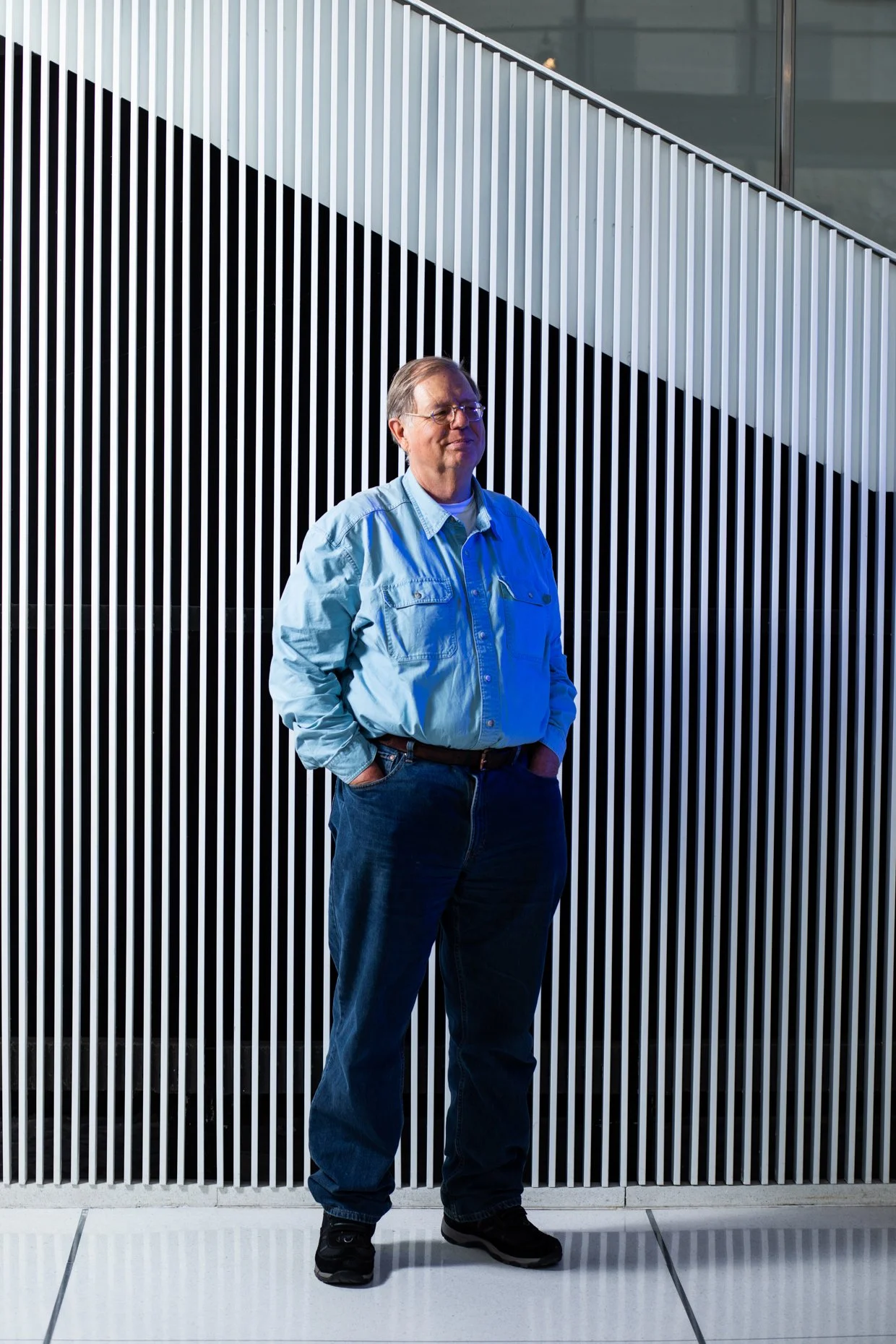 A man standing with his hands in his pockets in front of a black and white striped metal wall.