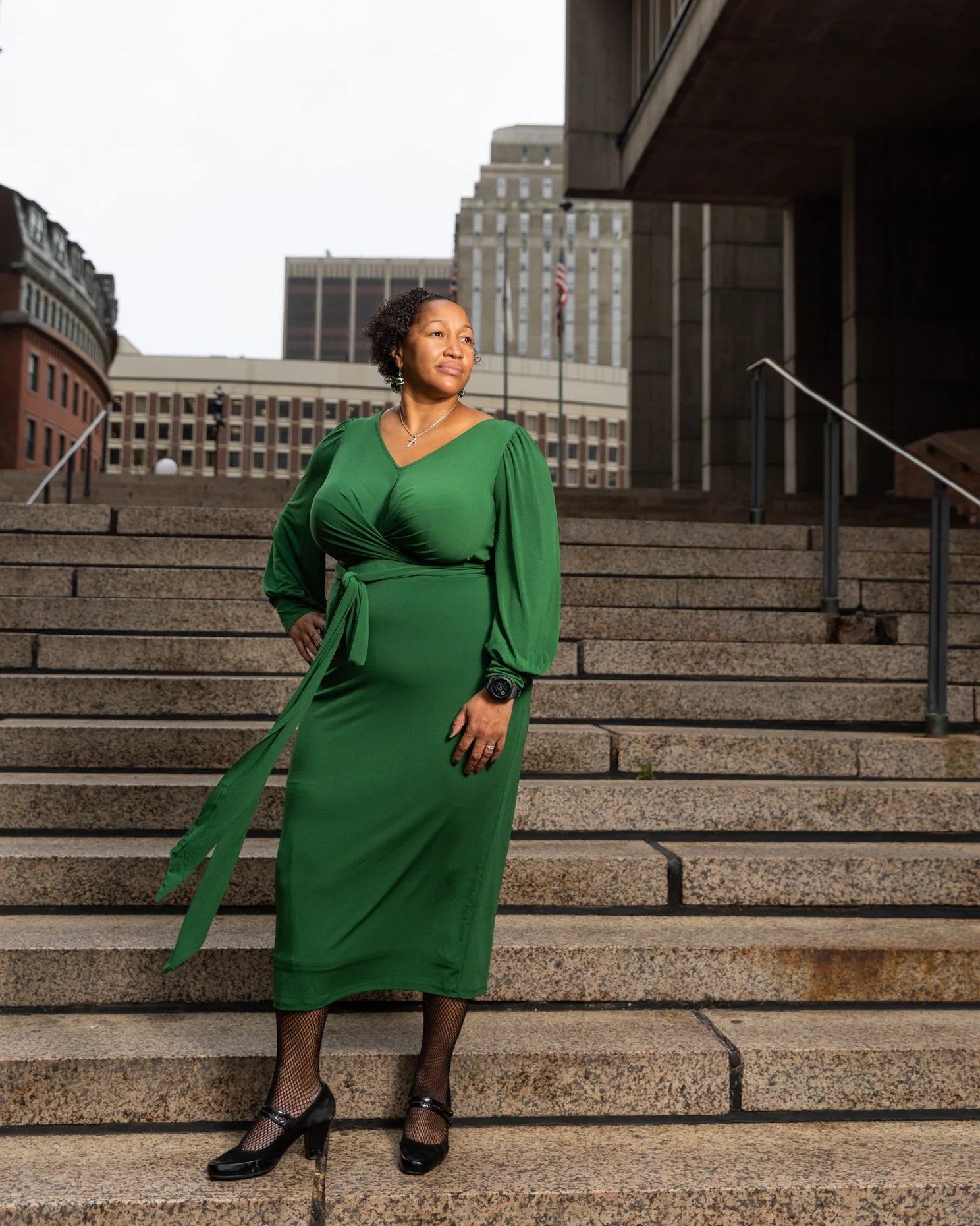 A woman in a long green dress with puffed sleeves and a tied waist, standing on outdoor stairs in an urban setting with tall buildings and American flags in the background. She is wearing fishnet stockings, high heels, and jewelry, looking off into t