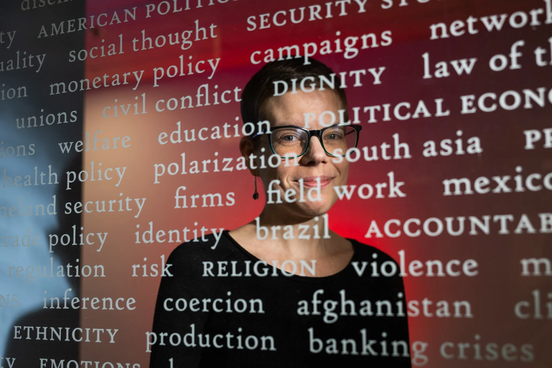 The image features a woman with short dark hair, wearing glasses, smiling at the camera. Behind her, there is a large word cloud with various words in white and light blue on a background that transitions from dark gray on the left to red on the righ