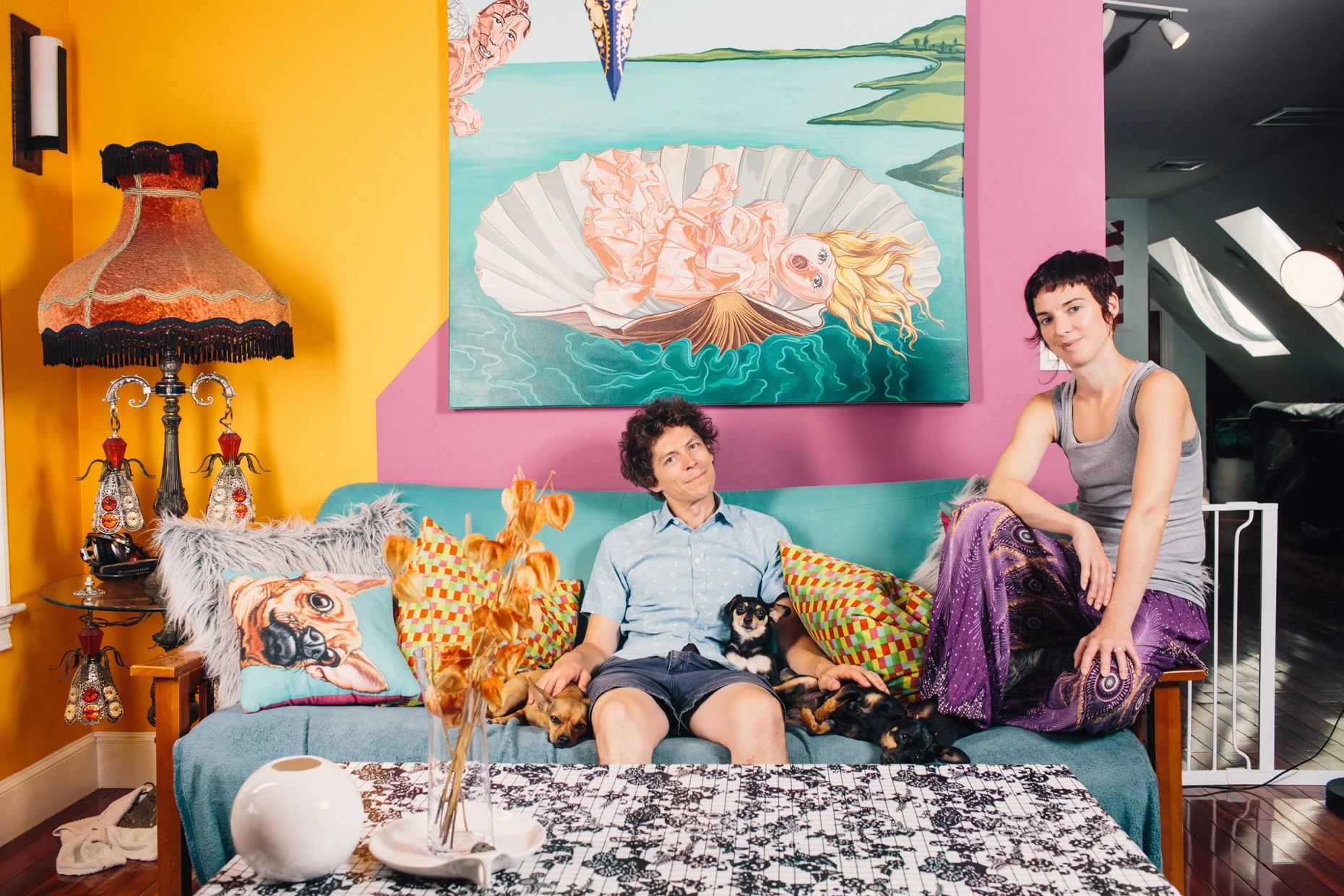 A colorful living room with two women, a man, and several dogs. The man is sitting on a blue couch with patterned pillows, and the woman with short dark hair stands beside him. The other woman has short dark hair and is seated on the arm of the couch