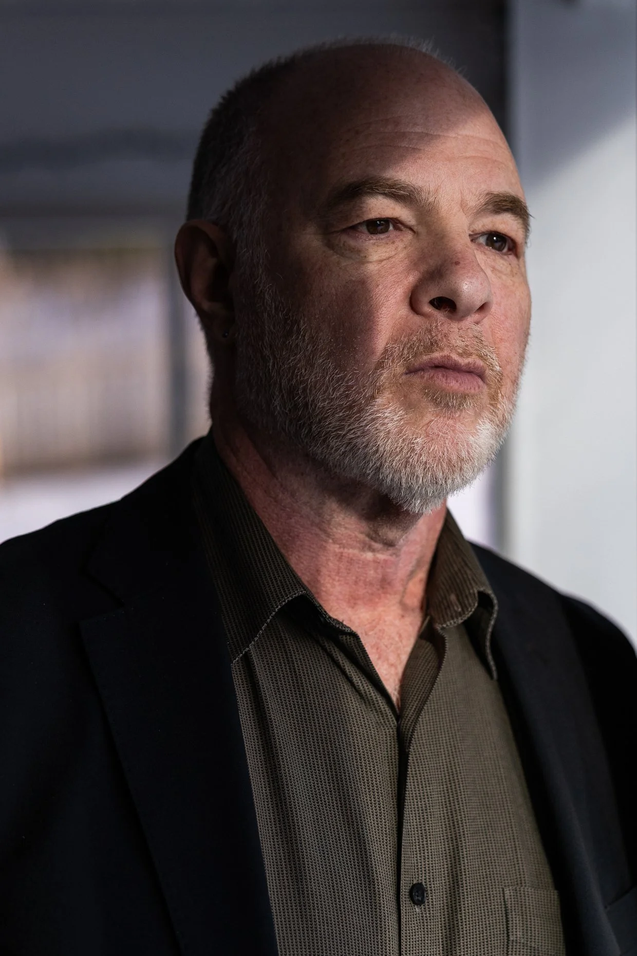 Close-up of a middle-aged man with a beard and short hair, wearing a black blazer and a brown shirt, standing indoors with a blurred background.