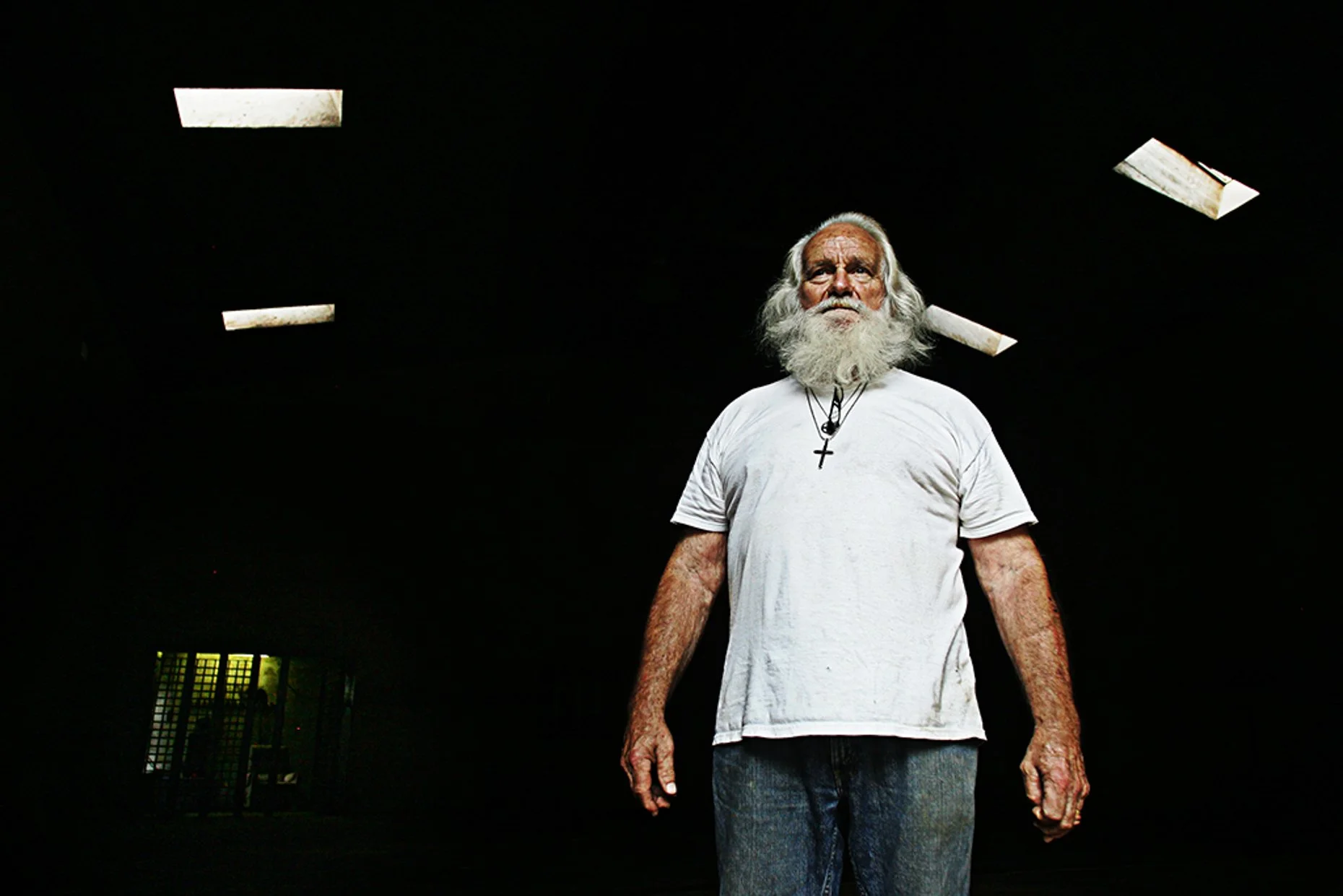 An elderly man with a long white beard and white hair, wearing a white t-shirt and a cross necklace, stands in a dark room with bright lights on the ceiling. There is a yellow and green lit window or LED display in the background.