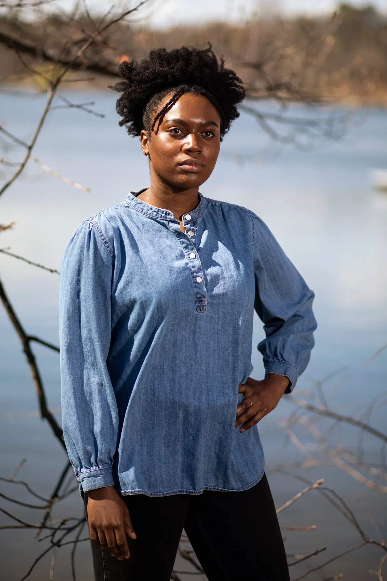 A woman standing outdoors near a body of water, wearing a long-sleeved denim shirt and black pants, with her right hand on her hip and looking directly at the camera.