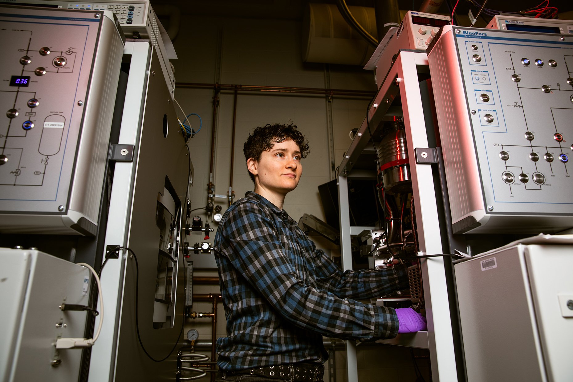 A woman working in a laboratory with scientific equipment and electronic devices, wearing a plaid shirt and purple gloves.