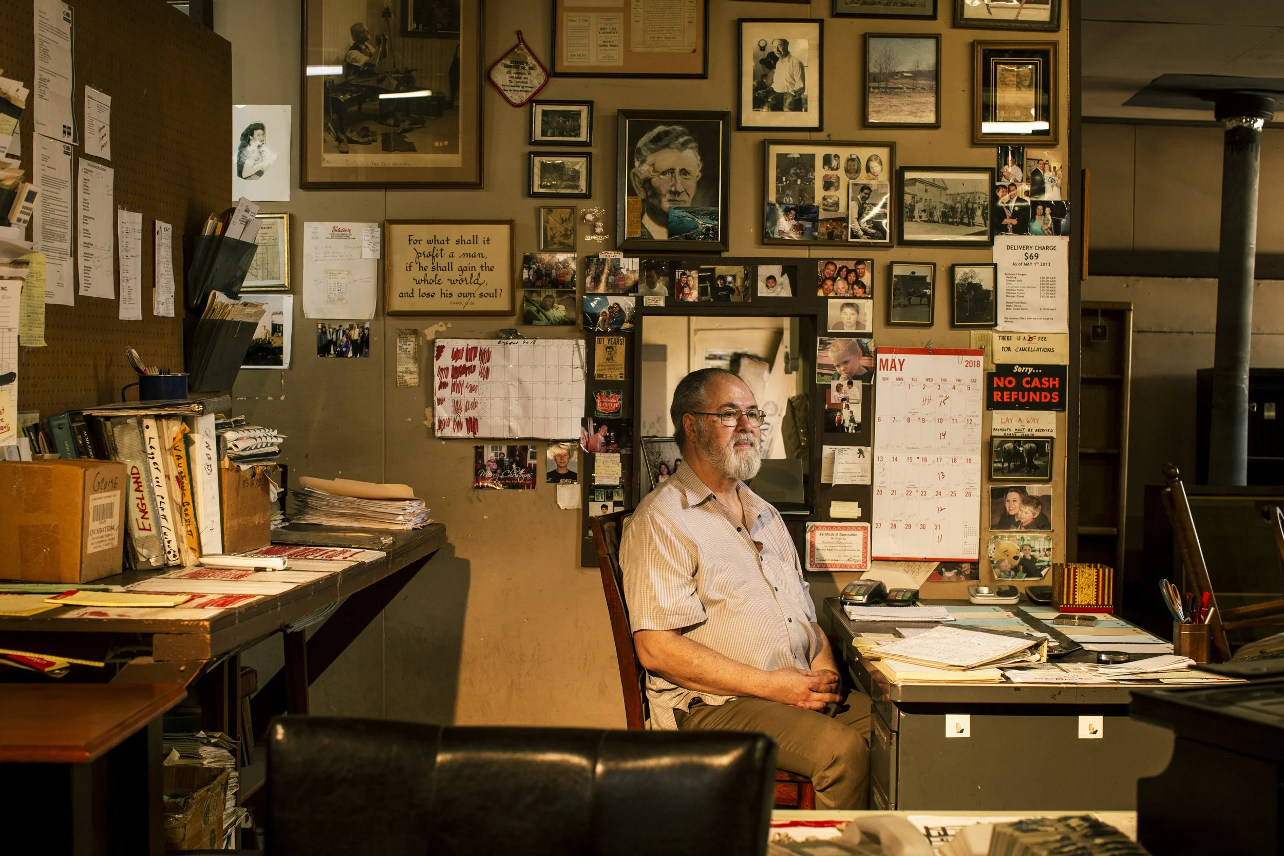 A man with glasses and a beard sitting in an office surrounded by numerous framed photos, newspaper clippings, and various papers on the walls and desk. The office has a cluttered, cozy feel with a calendar, sign, and a peghole board on the wall.