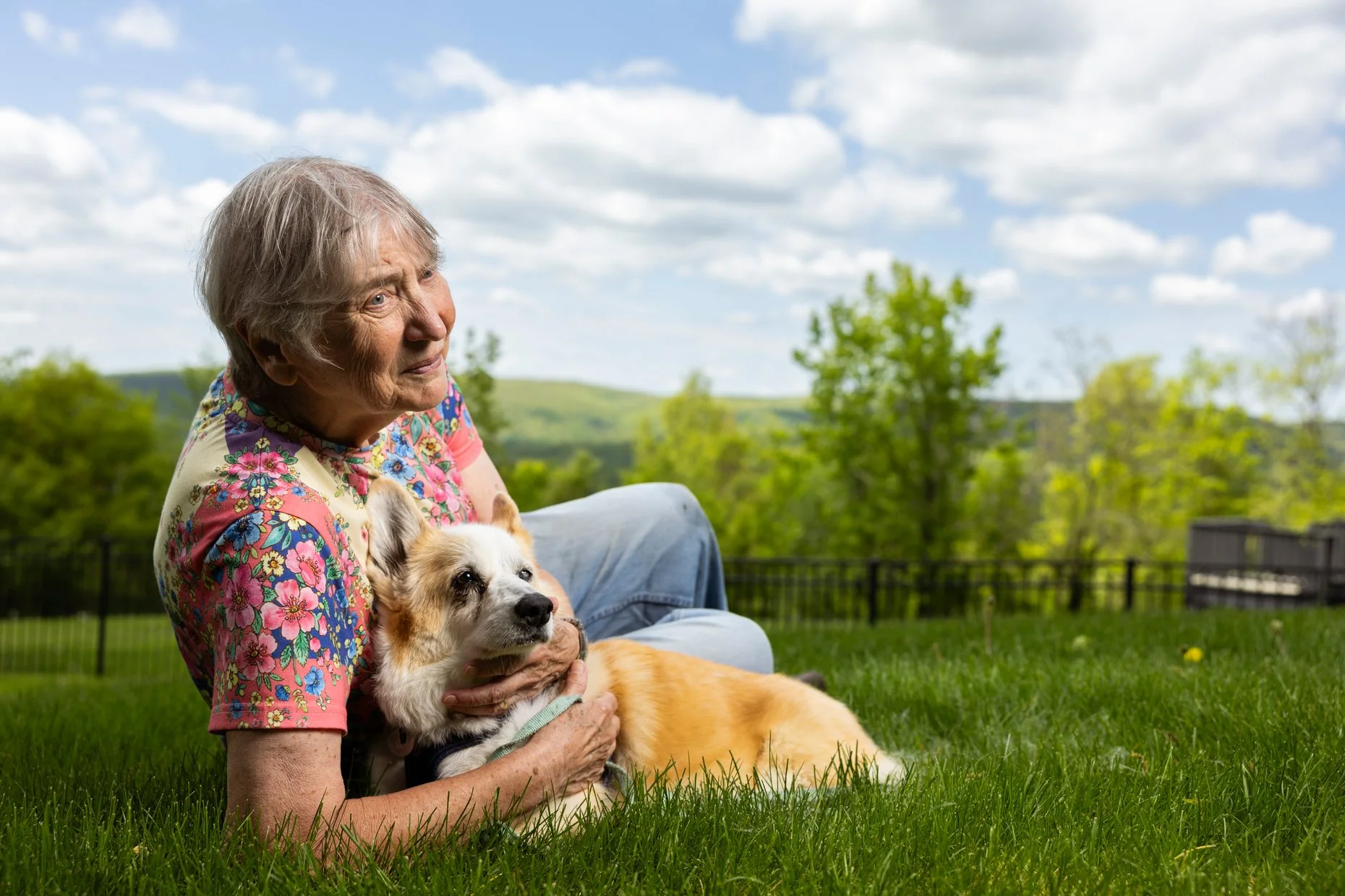 An elderly woman with gray hair and a colorful floral shirt is sitting on the grass outdoors, holding a small dog with white and tan fur. The woman looks into the distance, and the background features a blue sky with scattered white clouds, green tre