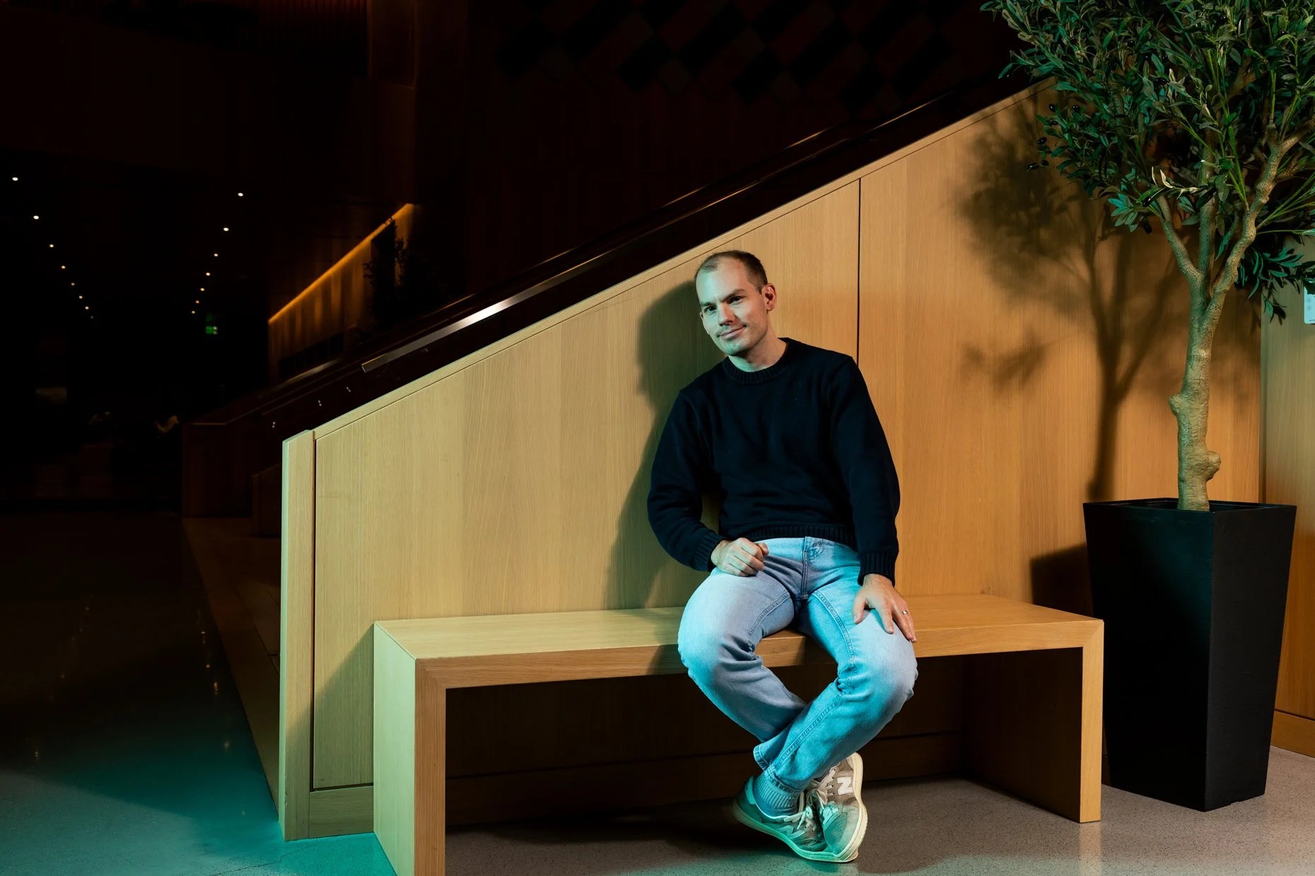 A man sitting on a wooden bench in a modern interior space with a large potted plant next to him, wearing a black sweater and light jeans.