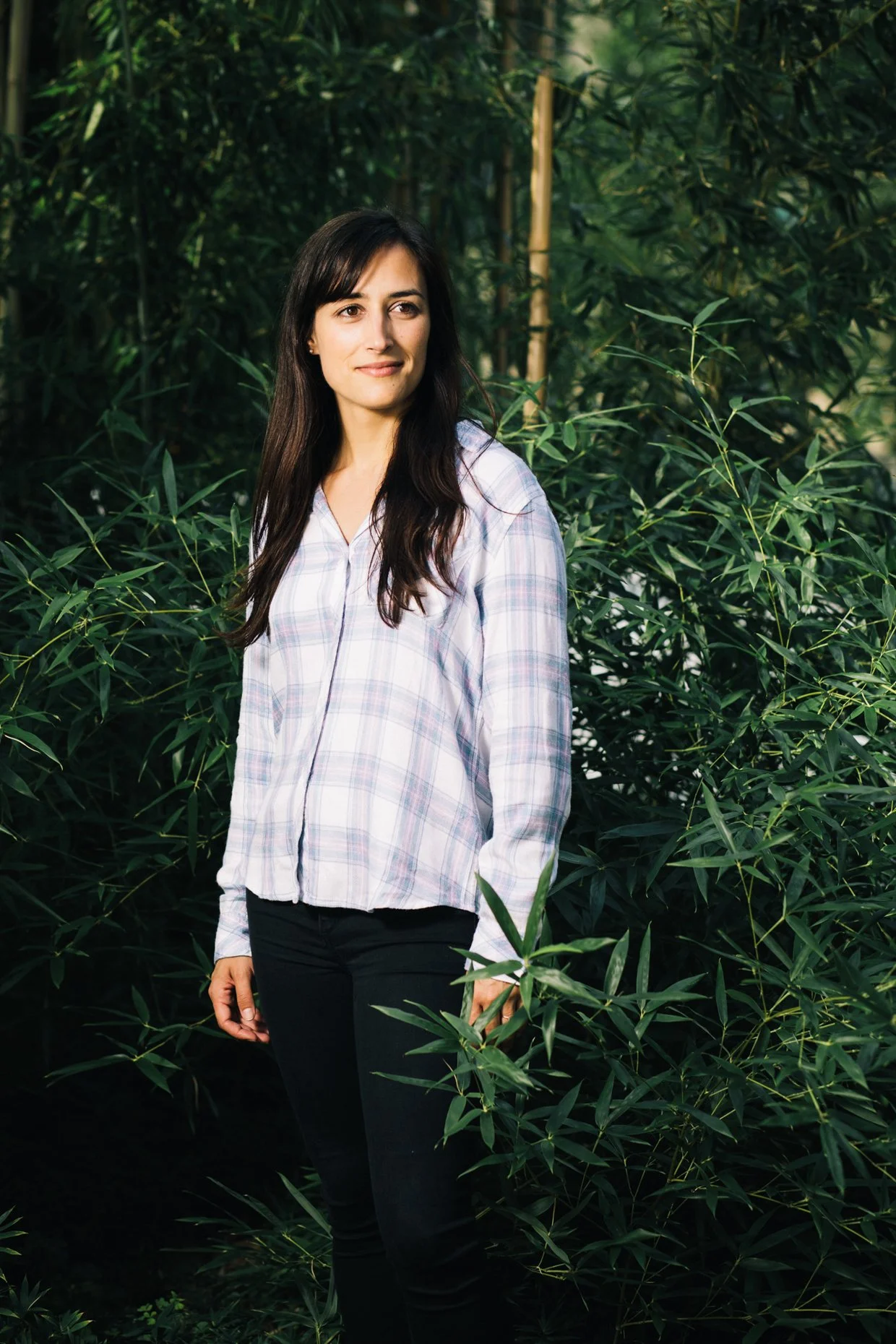 A woman with dark hair wearing a white plaid shirt and black pants standing outdoors among green leafy plants.
