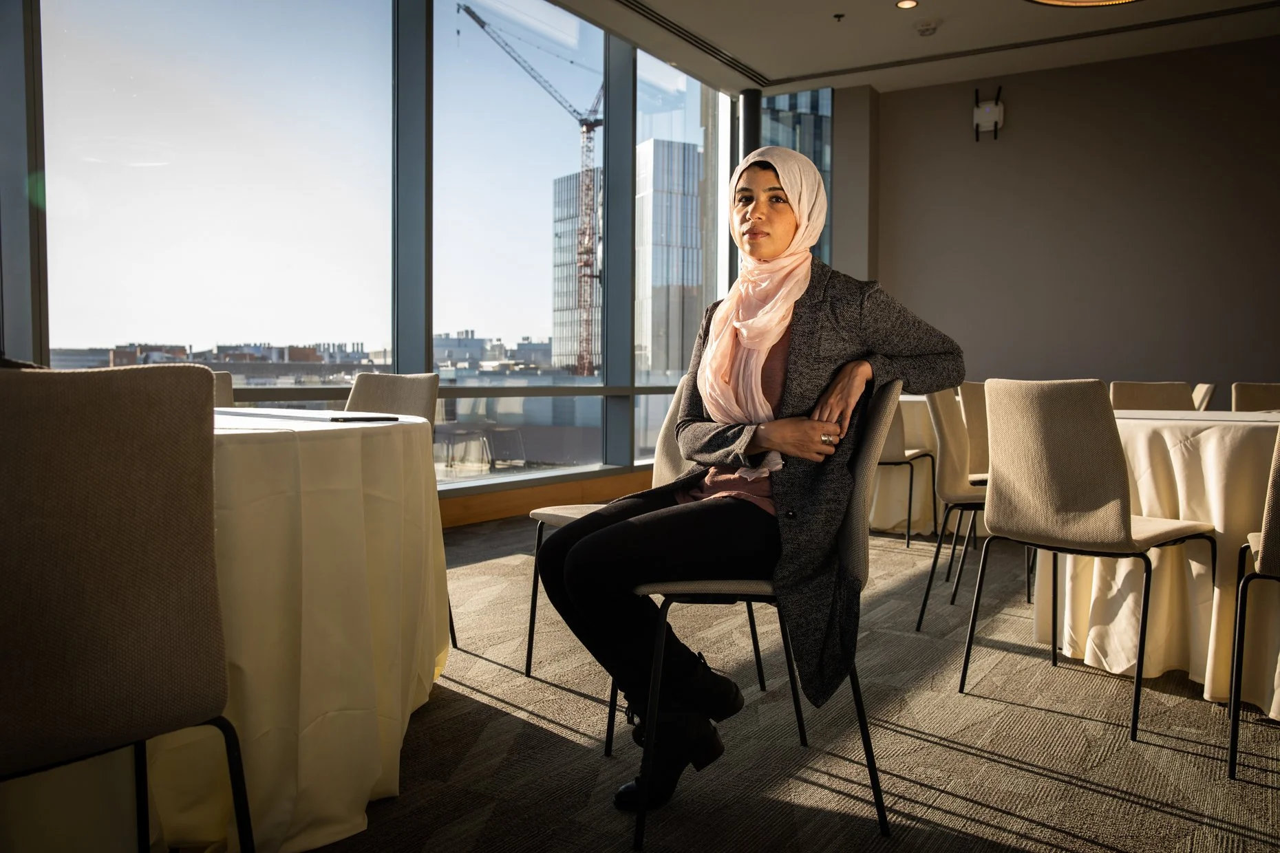 A woman wearing a light-colored hijab and a gray blazer, sitting in a conference room with large window views of a city and crane. She is seated at a table with yellow tablecloths, surrounded by beige chairs, with one arm resting on the chair's back.