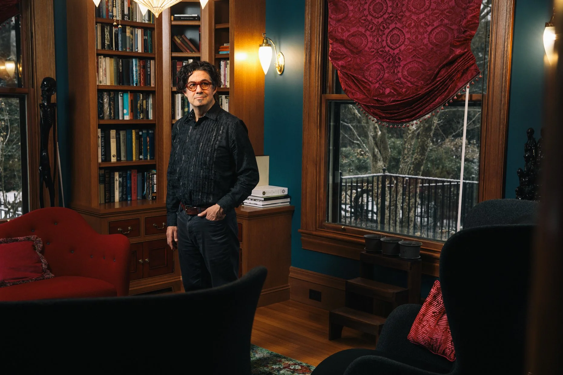 A man with dark, curly hair, wearing an orange pair of glasses, a black patterned shirt, and dark pants, stands inside a room with wooden trim and a large window with a red, patterned curtain. The room features a red velvet sofa, a wooden bookshelf f