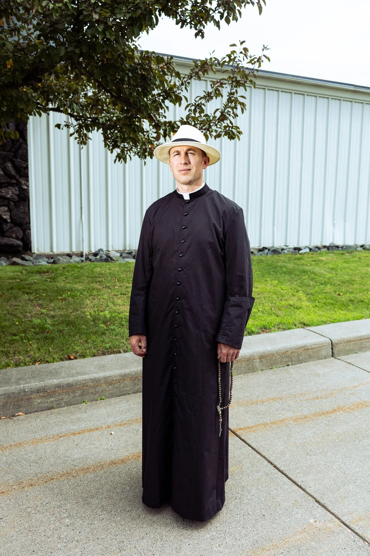 A man dressed as a priest, wearing a black cassock, a white clerical collar, and a white hat, stands on a sidewalk in front of a grassy area, a tree, and a building with white vertical siding.
