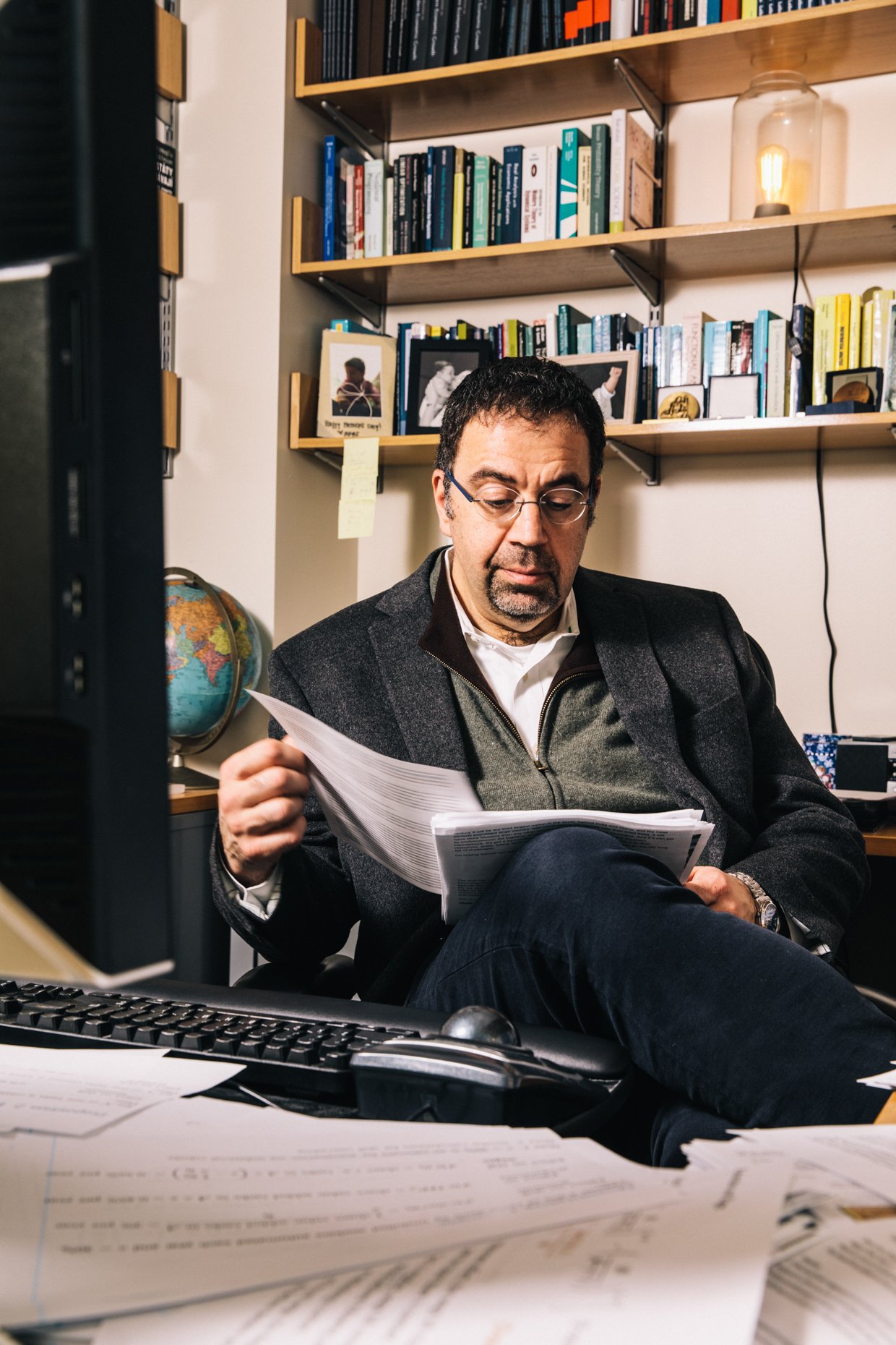 A man sitting at his desk in an office, looking at papers. Behind him are shelves filled with books, framed photos, and a glass lamp. The desk has scattered documents and a computer keyboard and mouse.