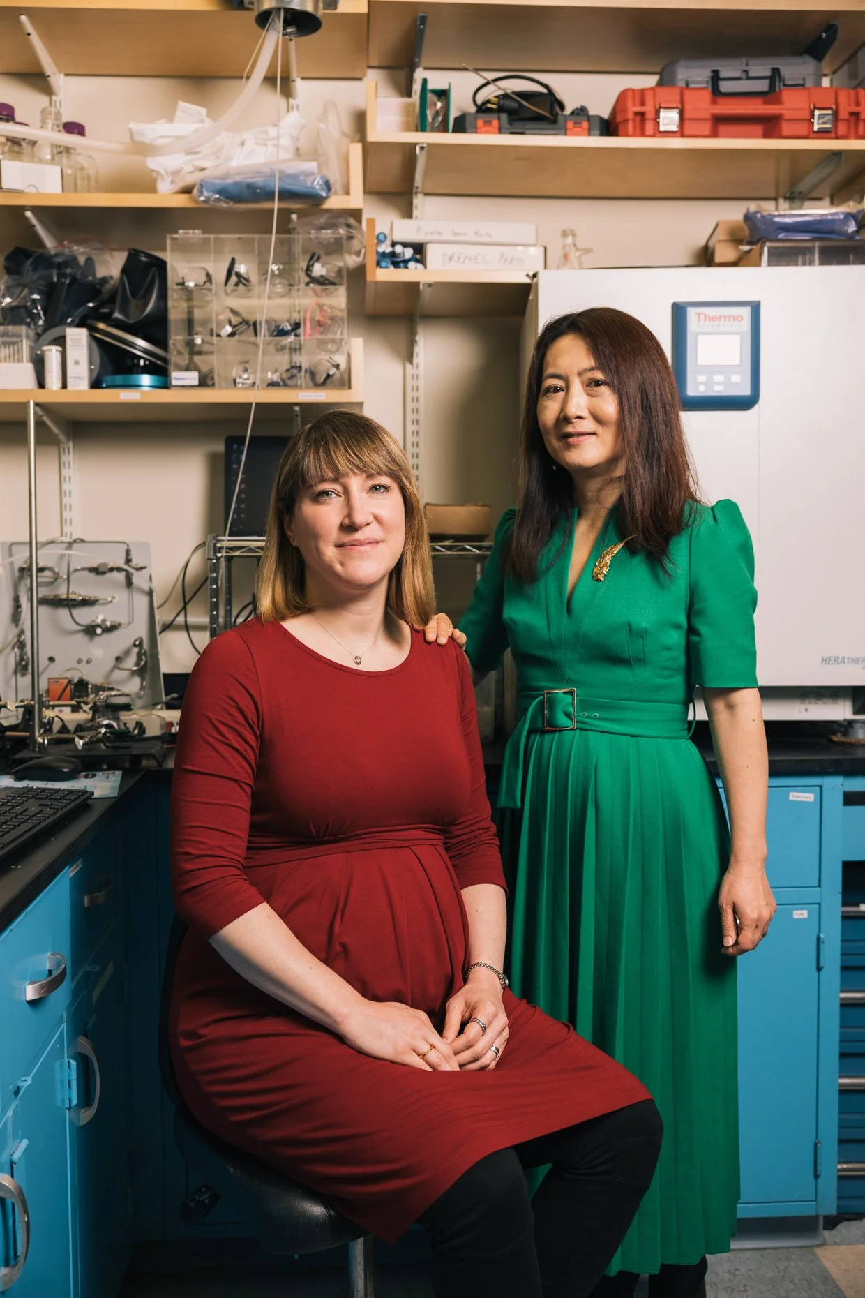 Two women in a laboratory, one seated and one standing, behind shelves filled with lab equipment and boxes.