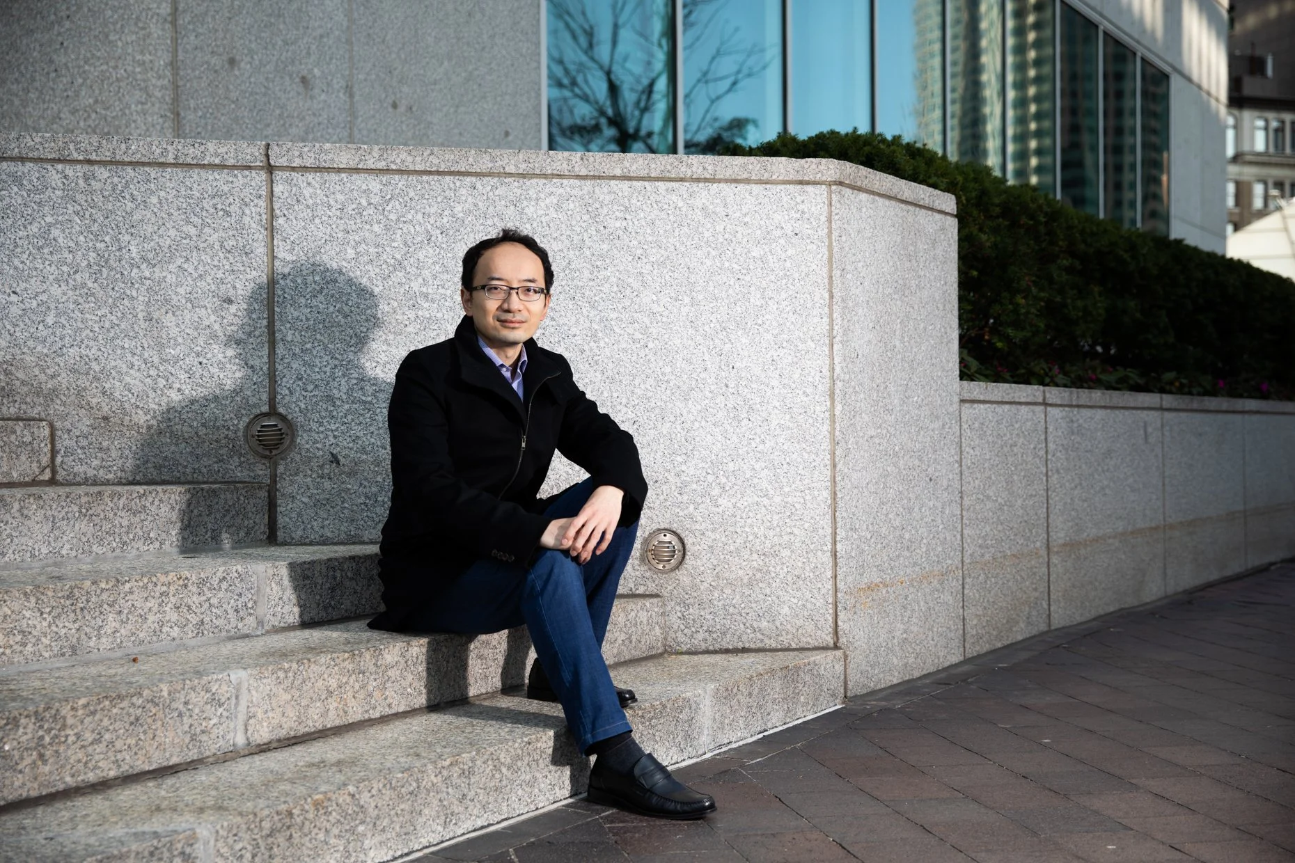A man sitting on stone steps outdoors, wearing glasses, a black jacket, and blue jeans, with glass buildings and trees reflected in the background windows.