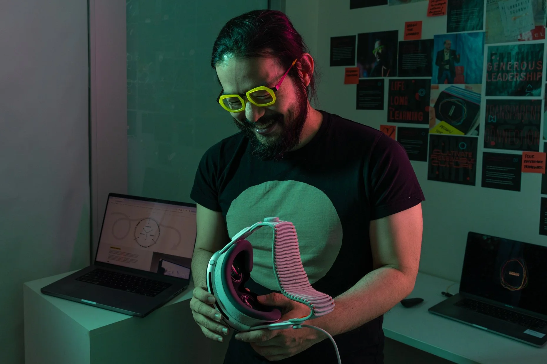 Man with long dark hair and beard, wearing yellow sunglasses and a black T-shirt, smiling while holding a pair of white headphones. In the background, there are posters on the wall and a laptop on a white table.