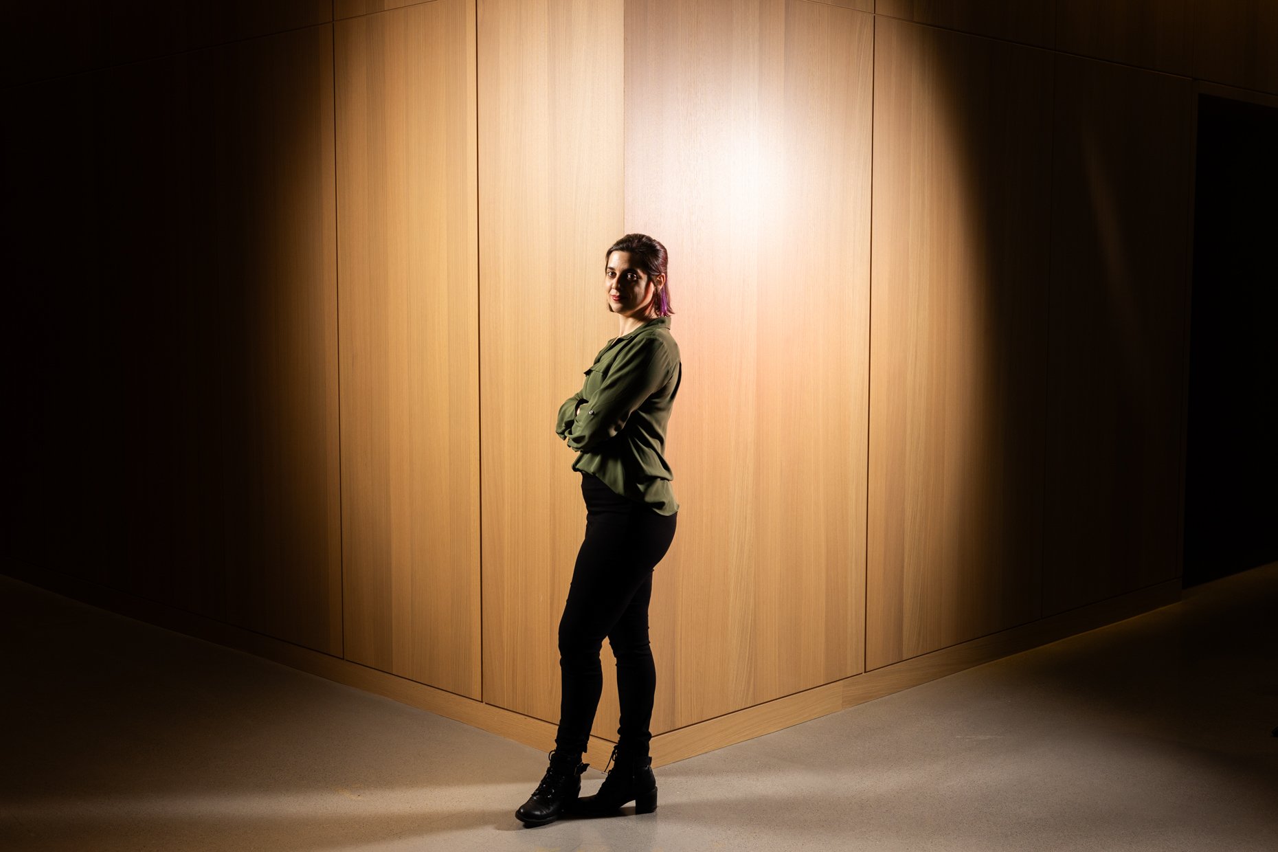 A woman with dark hair tied back, wearing a green jacket, black pants, and black boots, is standing with crossed arms in front of a wooden wall with spotlight lighting overhead, creating a circular light pattern around her.