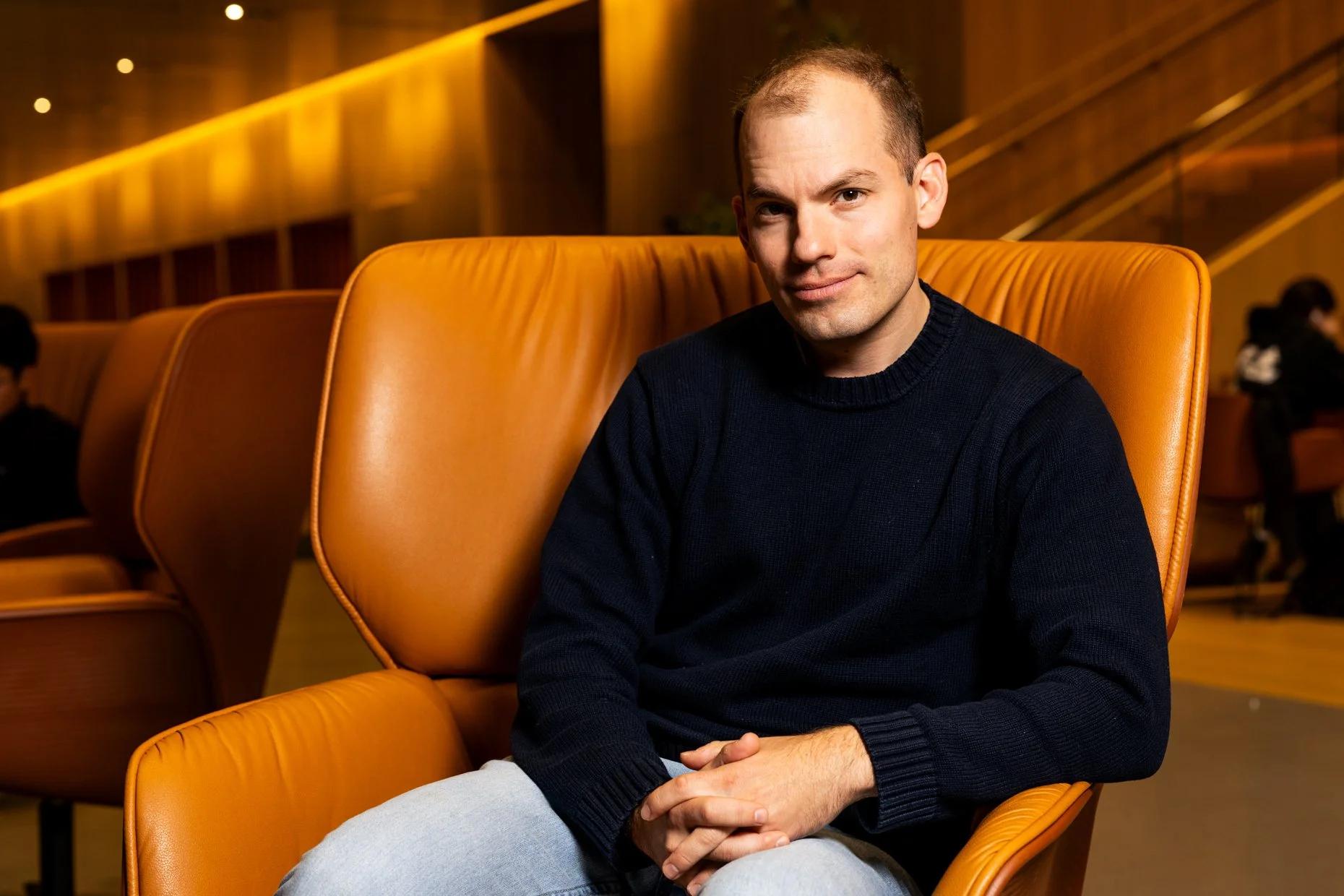 A young man with a buzz cut hairstyle, wearing a black sweater and light jeans, sitting in an orange leather chair in a warmly lit theater or auditorium, with rows of similar orange chairs behind him and a staircase in the background.