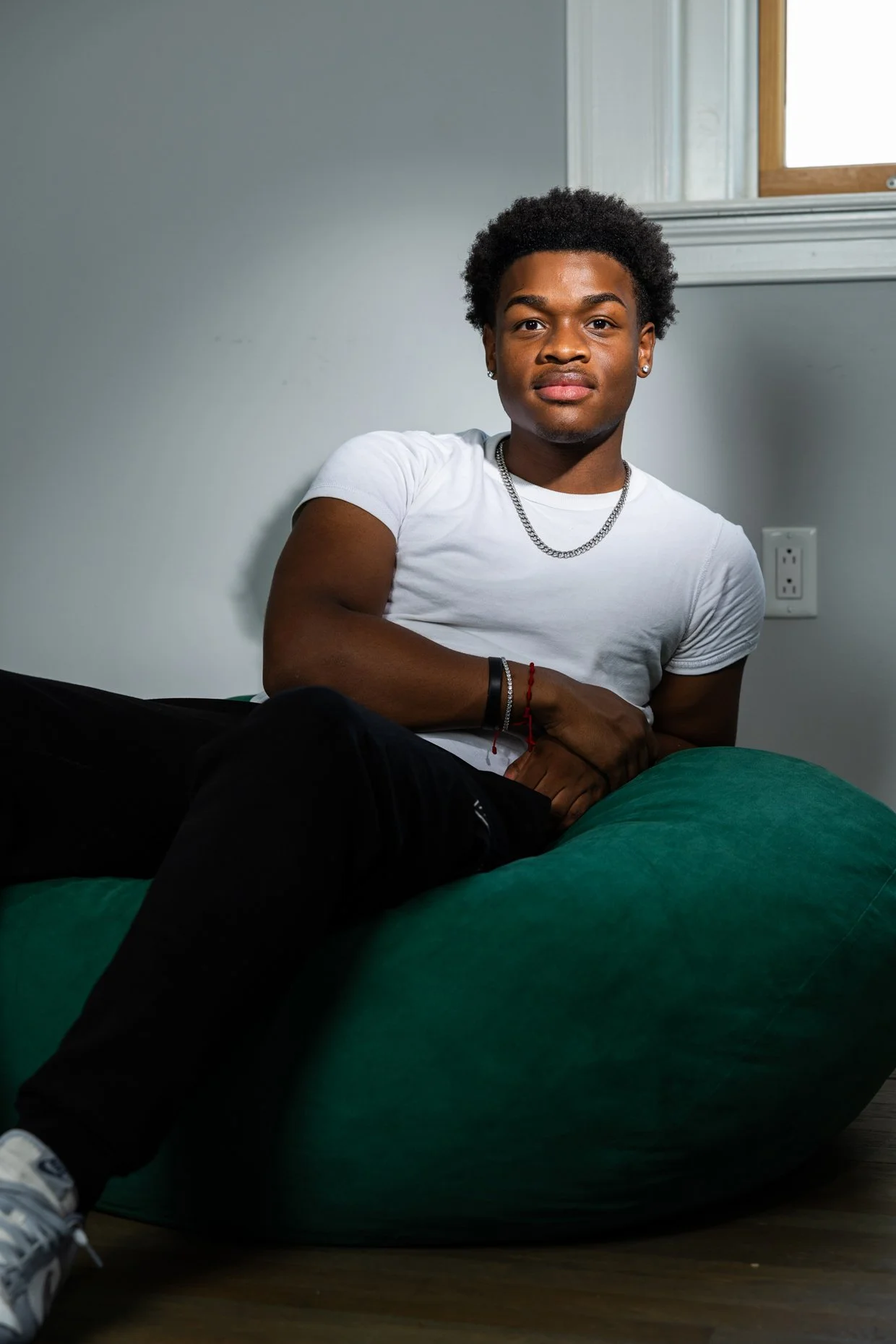 Young man with curly hair, wearing a white t-shirt, sitting on a green bean bag, and looking at the camera in a room with a plain wall and a window.