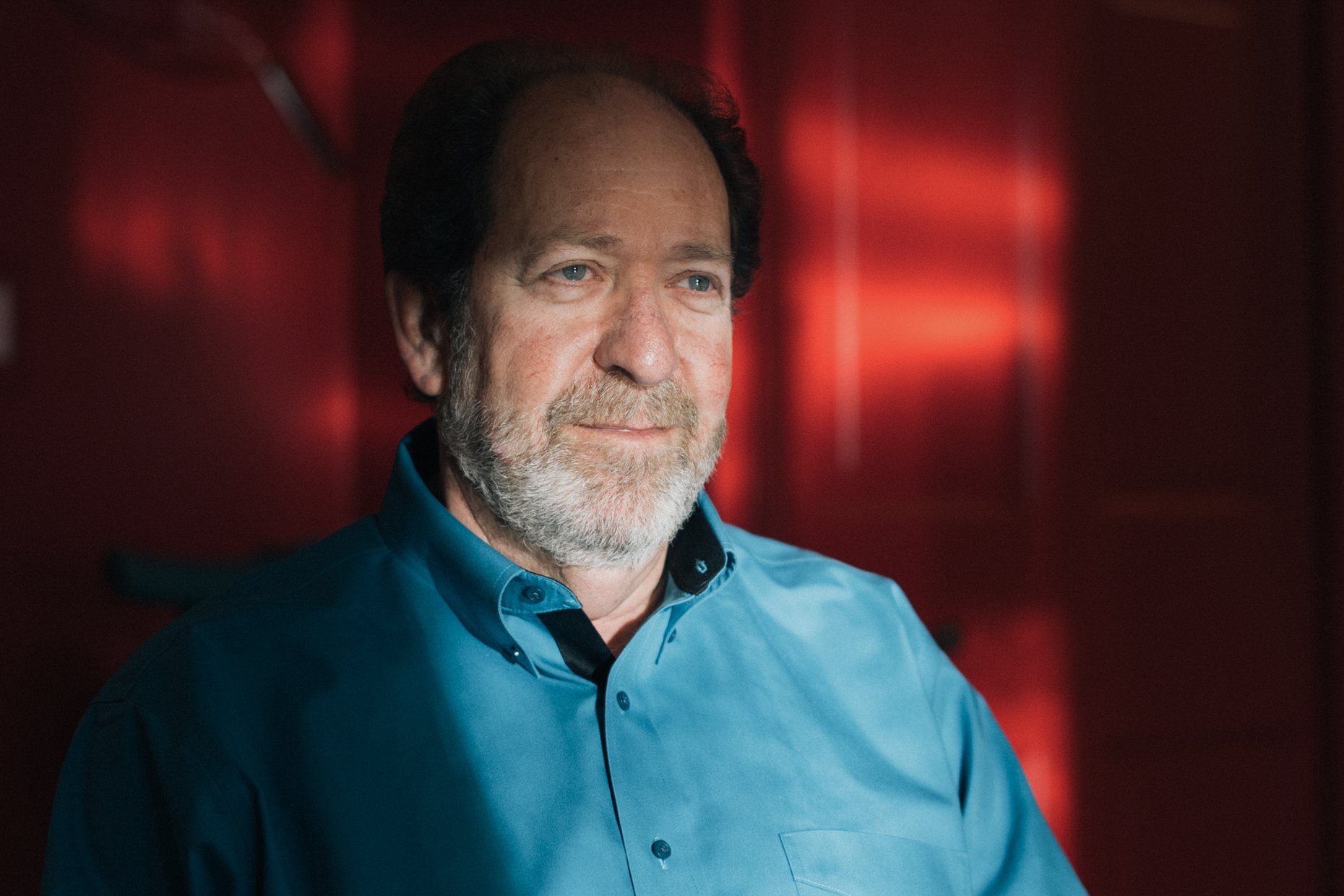 A middle-aged man with gray hair and a beard, wearing a blue button-up shirt, sitting in a room with a dark red background.