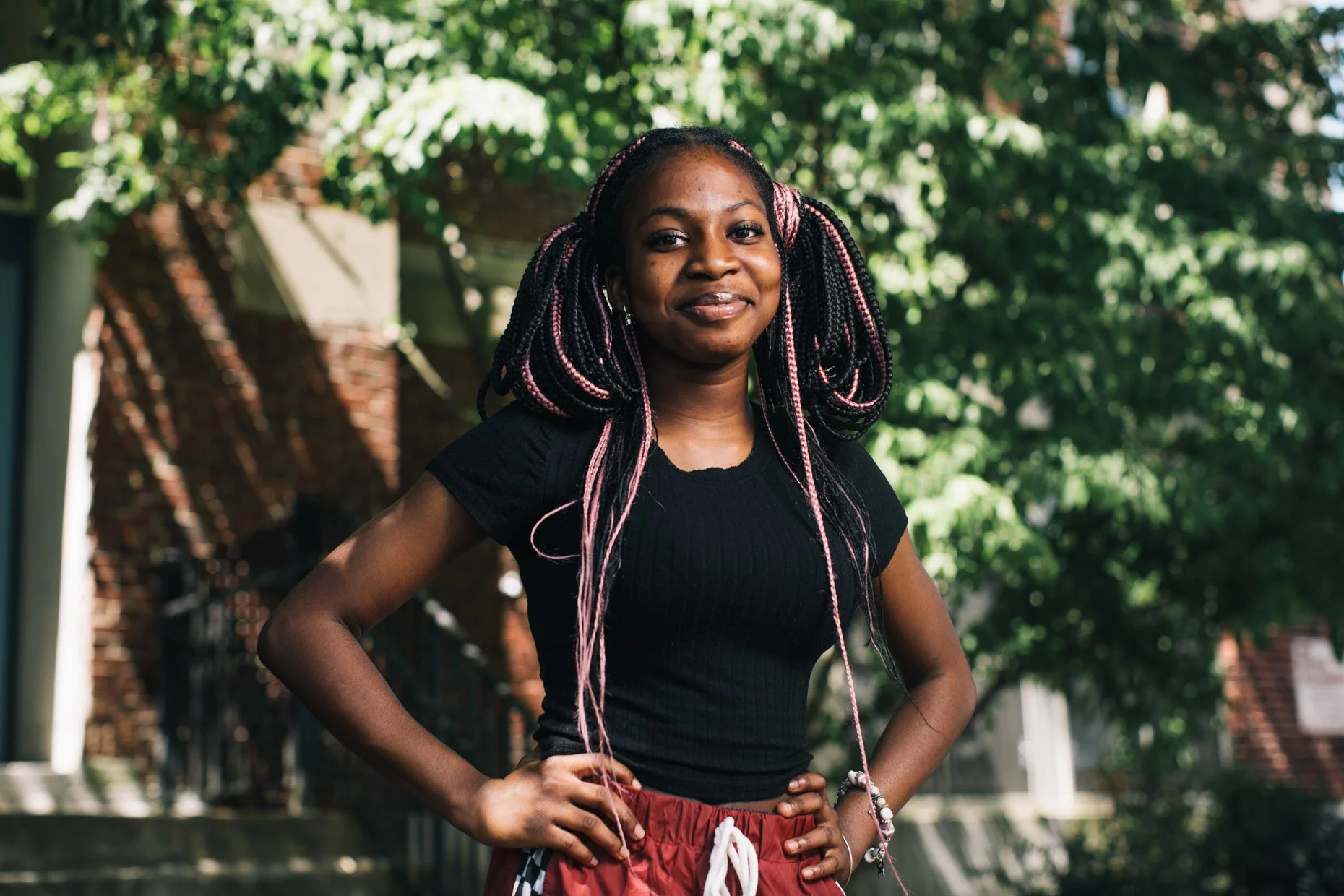 A young woman with braided hair with pink highlights, wearing a black T-shirt and red shorts, standing outdoors with her hands on her hips and smiling at the camera. Green trees and a brick building are in the background.