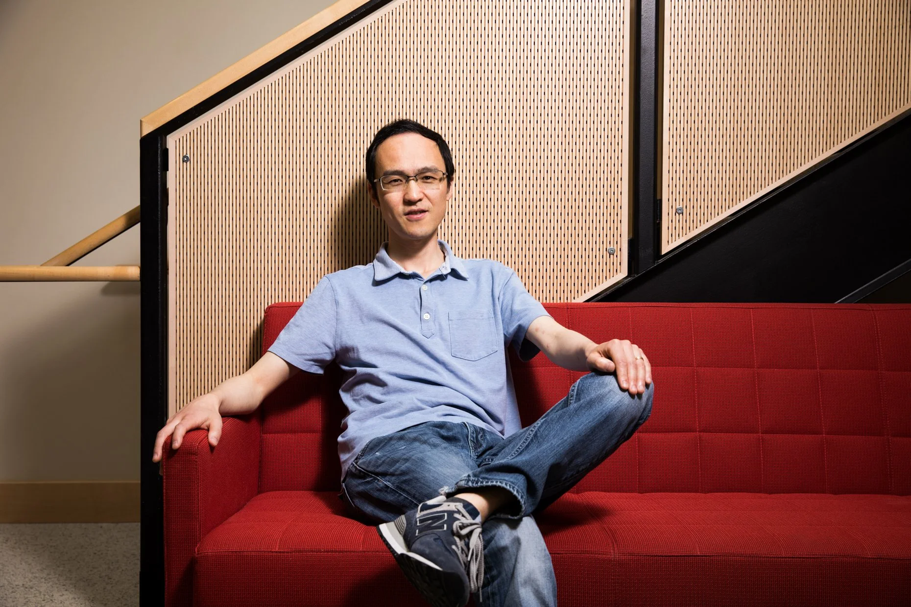 A man in a light blue polo shirt and jeans sitting on a red couch, with one leg crossed over the other, in an indoor setting with wooden wall panels behind him.