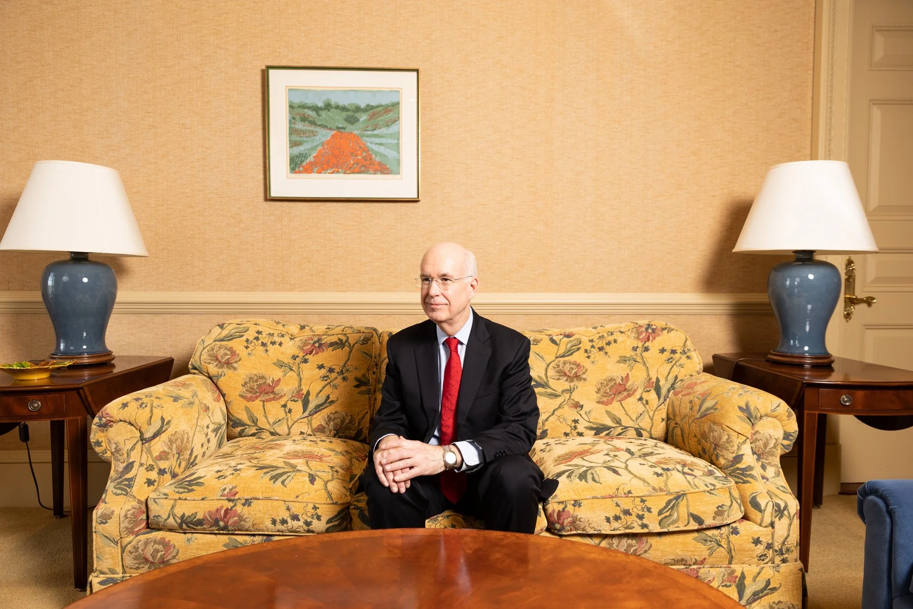 A man in a black suit, white shirt, and red tie sitting on a floral-patterned yellow sofa in a room with beige walls. There are two blue lamps on dark wood side tables on each side of the sofa. A framed landscape painting hangs on the wall behind him