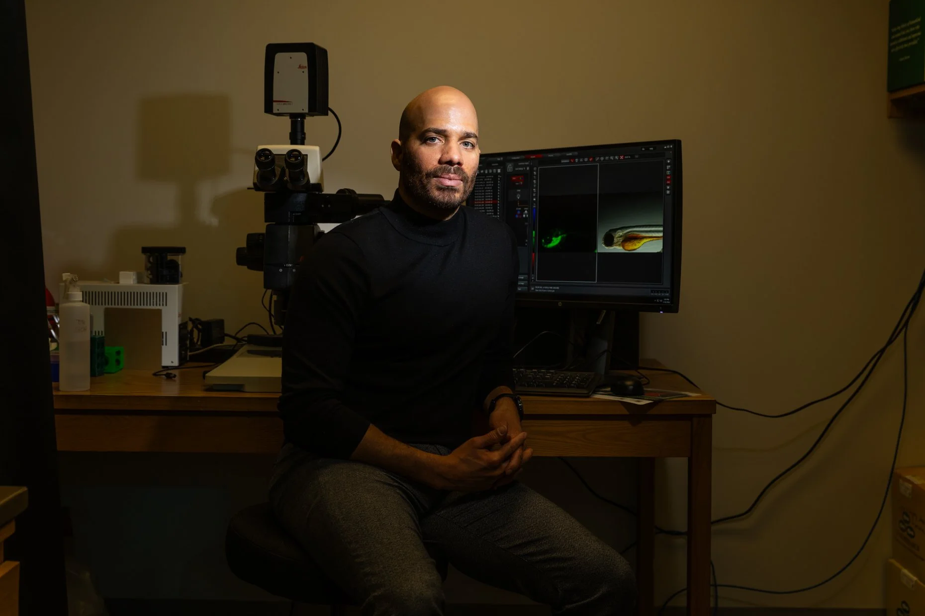 A man sitting at a desk with scientific equipment and a computer monitor displaying microscopy images, in a laboratory setting.