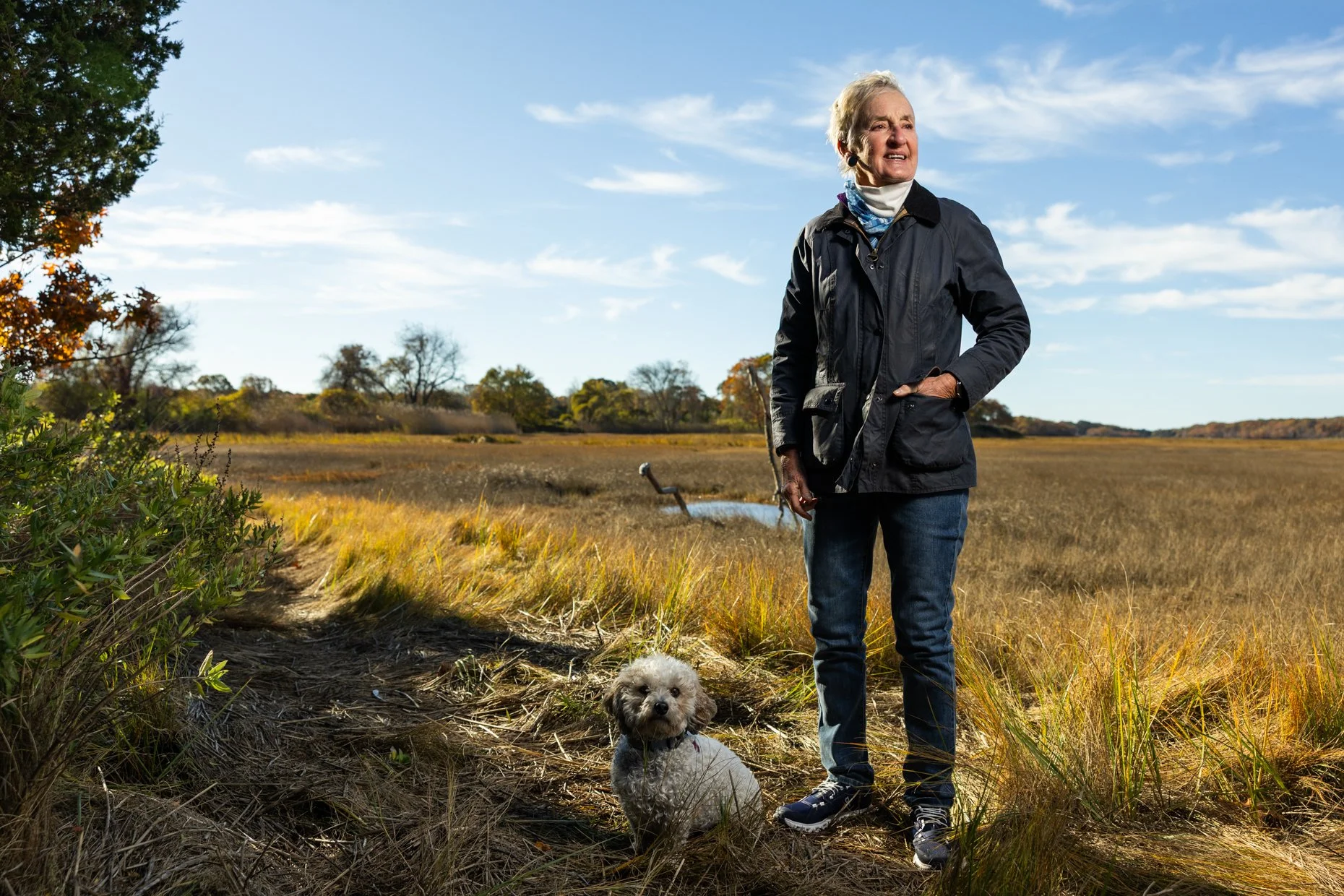 An elderly woman with short gray hair wearing a black jacket, jeans, and sneakers, standing outdoors in a field on a sunny day. She has a small dog sitting on the ground in front of her, and she is looking off to the side. The background features a w