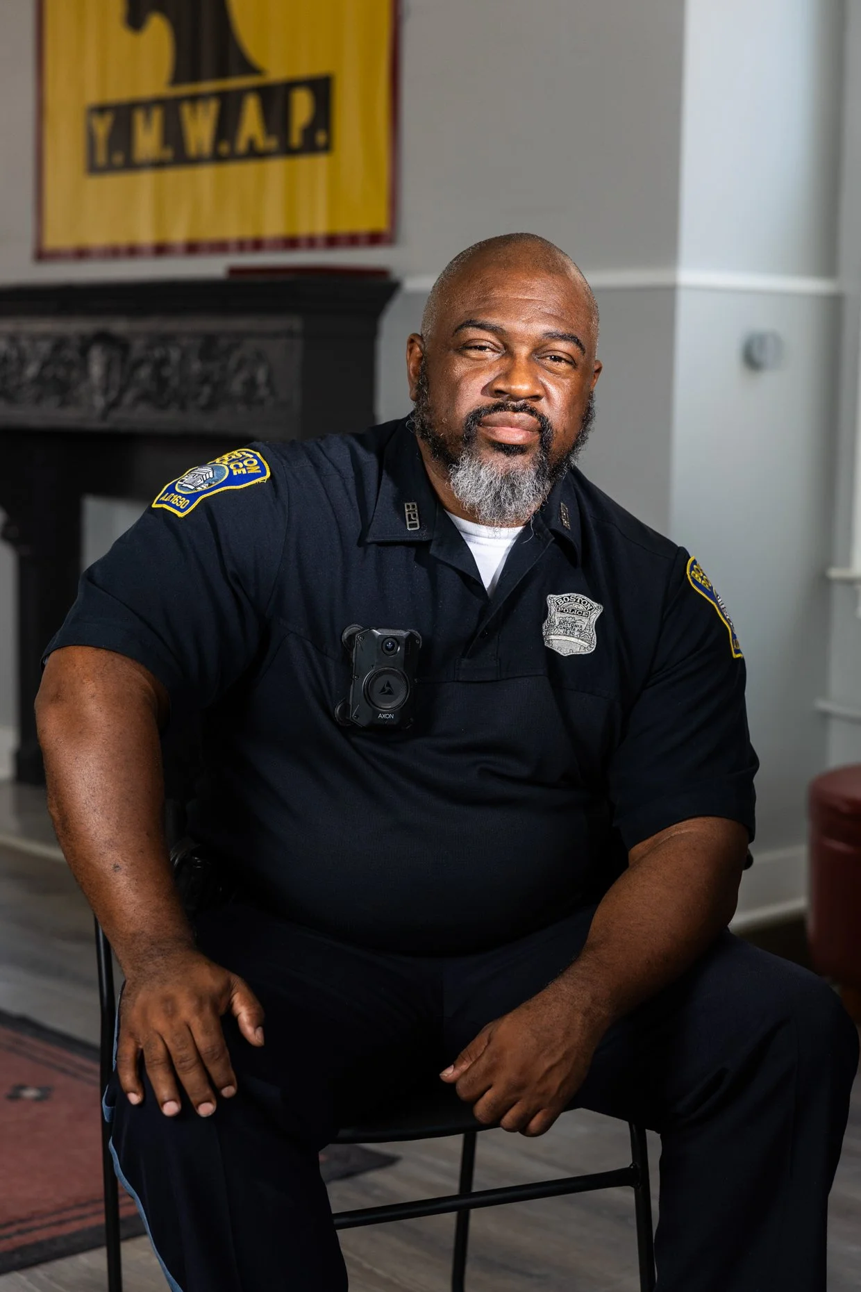 A bald African American police officer with a gray beard, wearing a dark uniform with patches and a badge, sitting in a room with white walls and a yellow banner in the background that reads 'Y.M.W.A.P.'