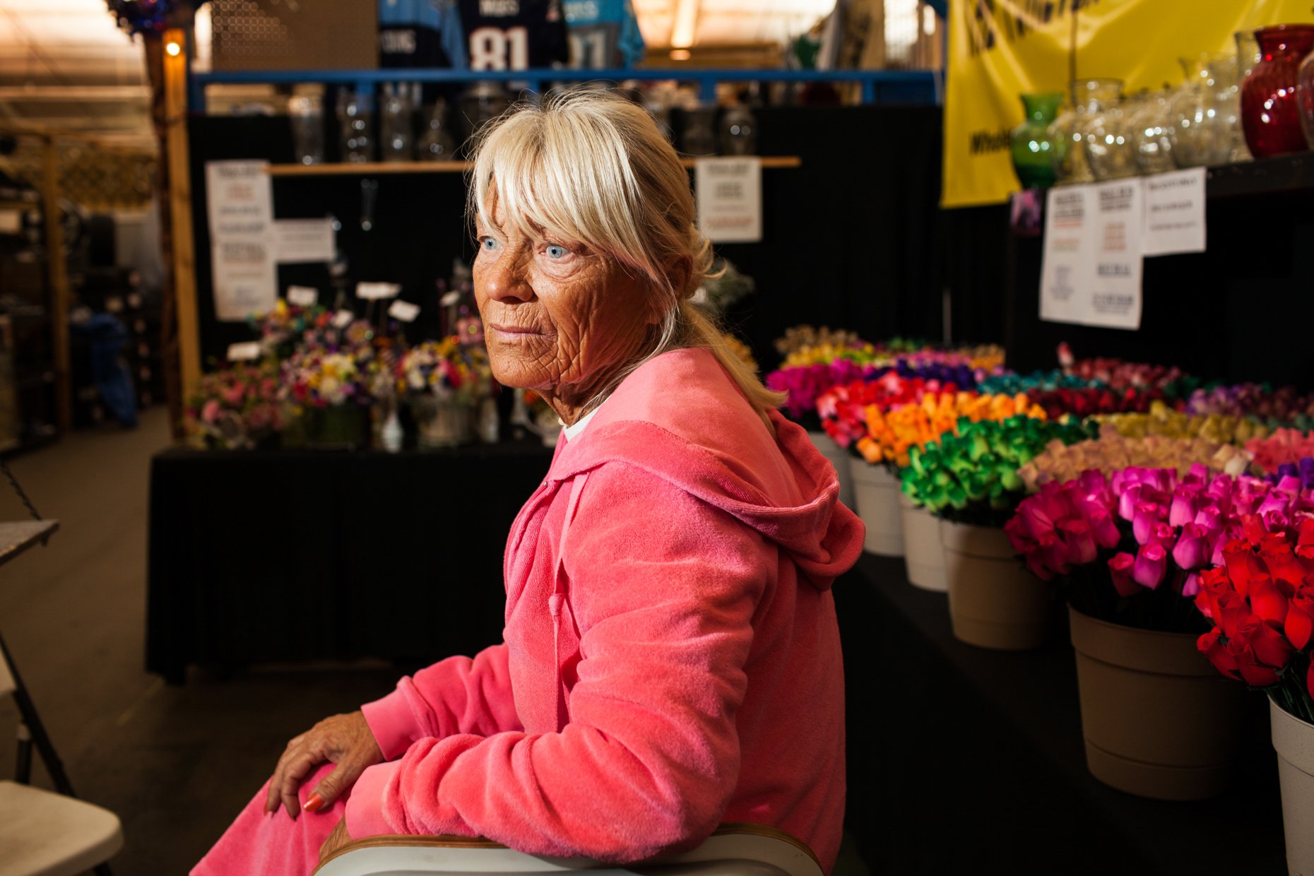 An elderly woman with blonde hair and blue eyes sitting among colorful flowers at a flower stand in a market or store.